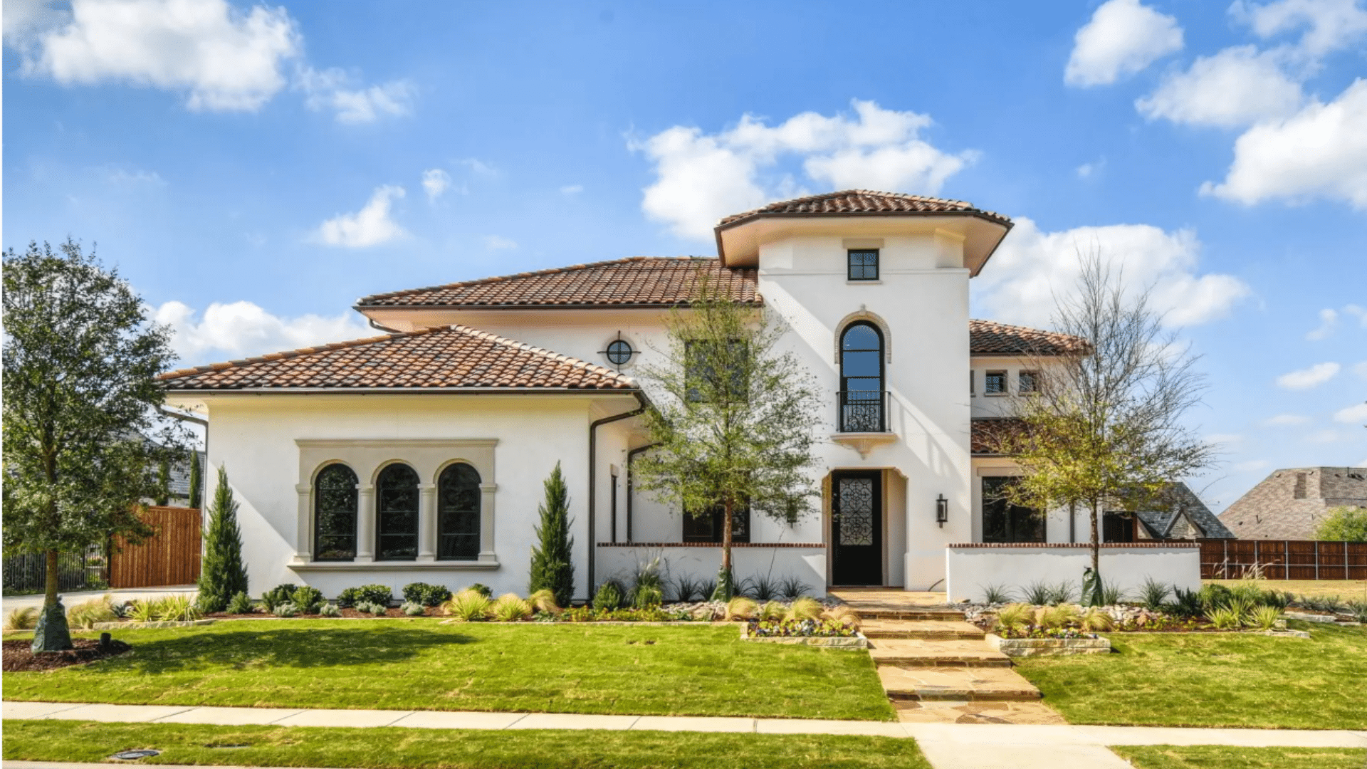 A Mediterranean style white house with a brown tile roof and arched windows on a green lawn under a blue sky with clouds