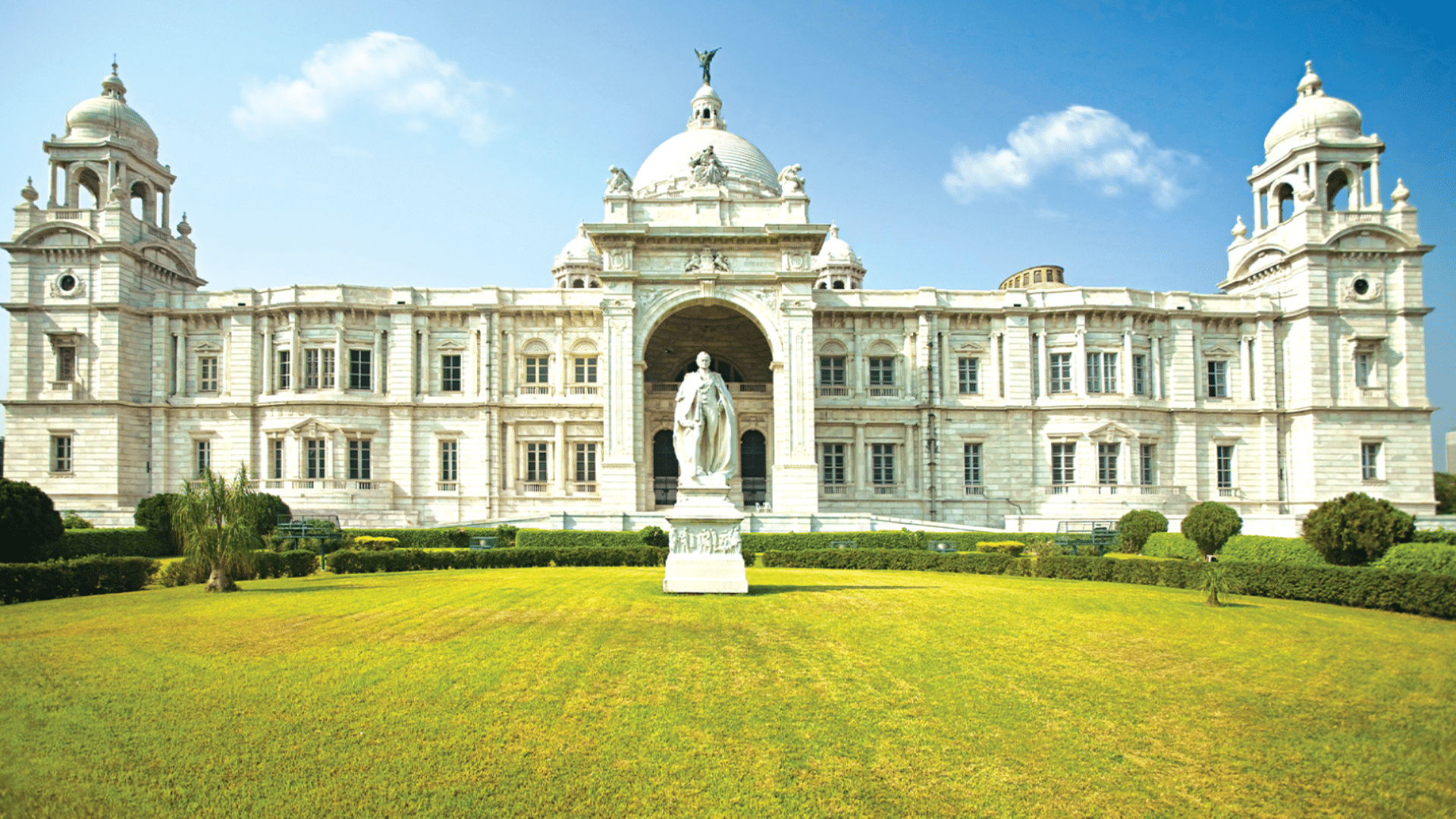 A large white marble monument with three high arched walkways stands on a stone base under a clear blue sky in the day