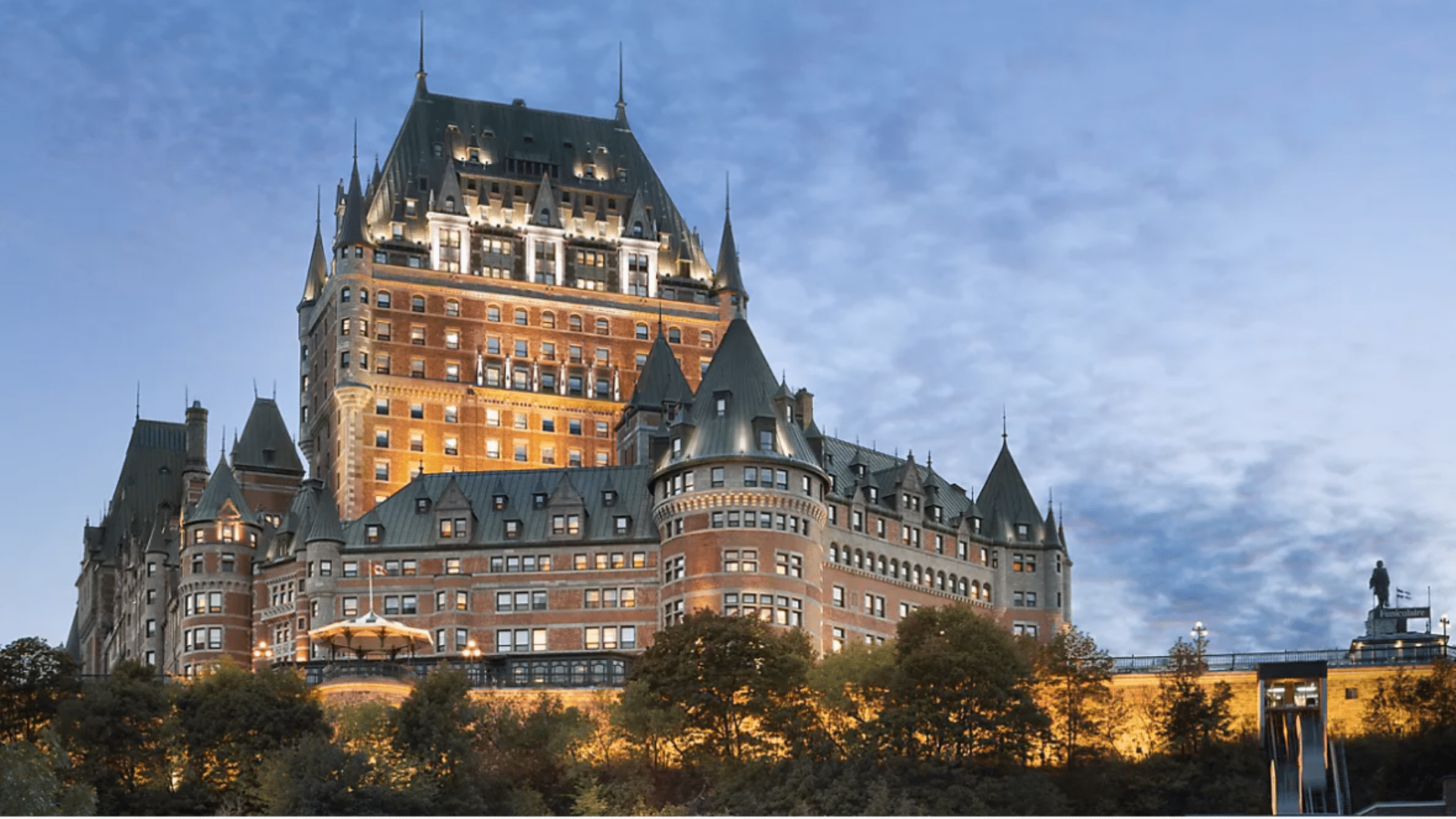 Grand brick hotel with many lit windows and green pointed towers stands on a hill against a blue evening sky with clouds