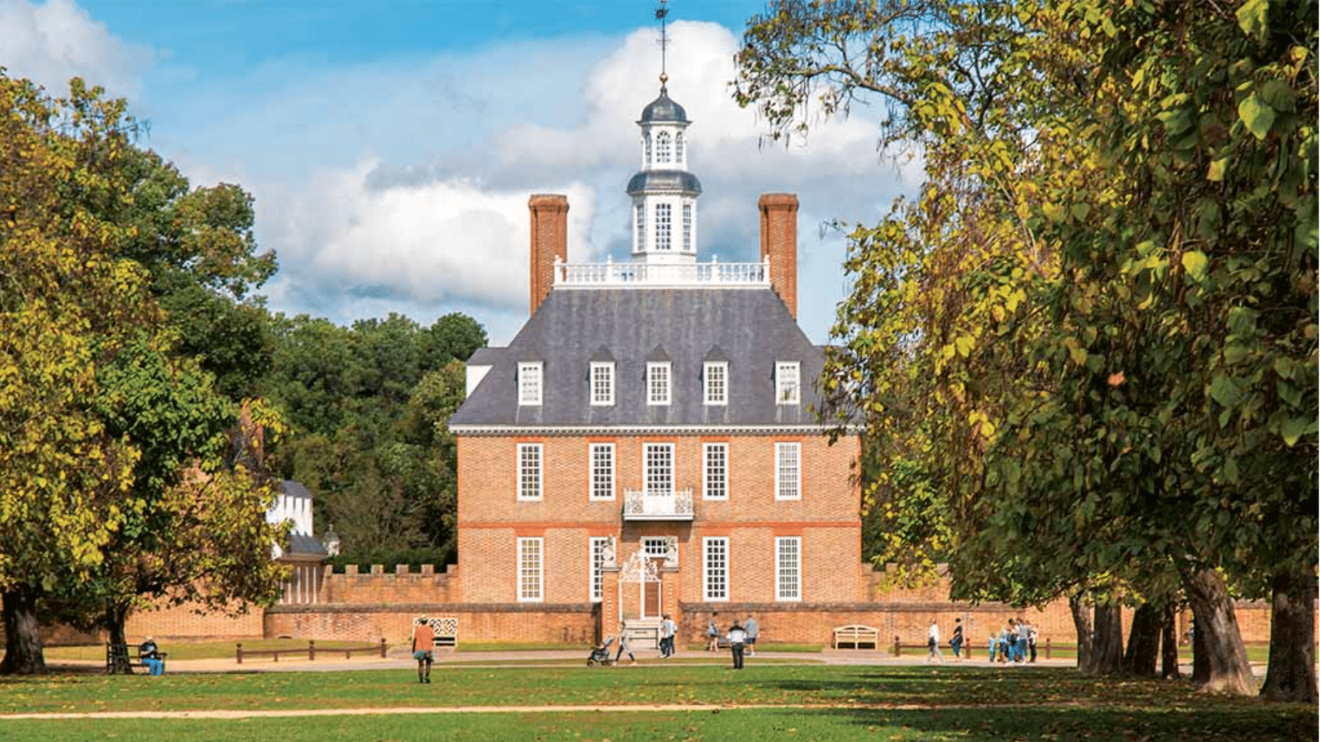 Large brick building with a white tower and two tall chimneys stands on a green lawn with leafy trees and people walking