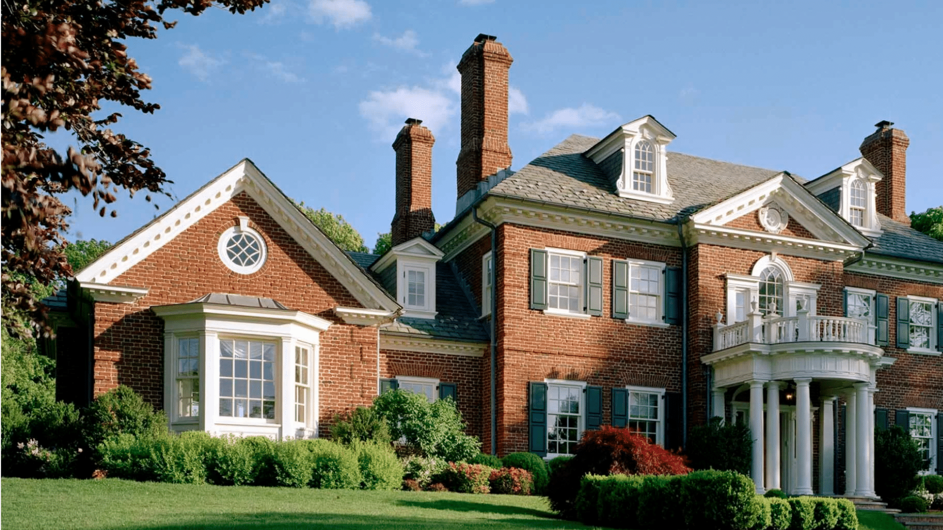 Large red brick mansion with white pillars, green shutters, and tall chimneys on a grassy hill under a bright blue sky