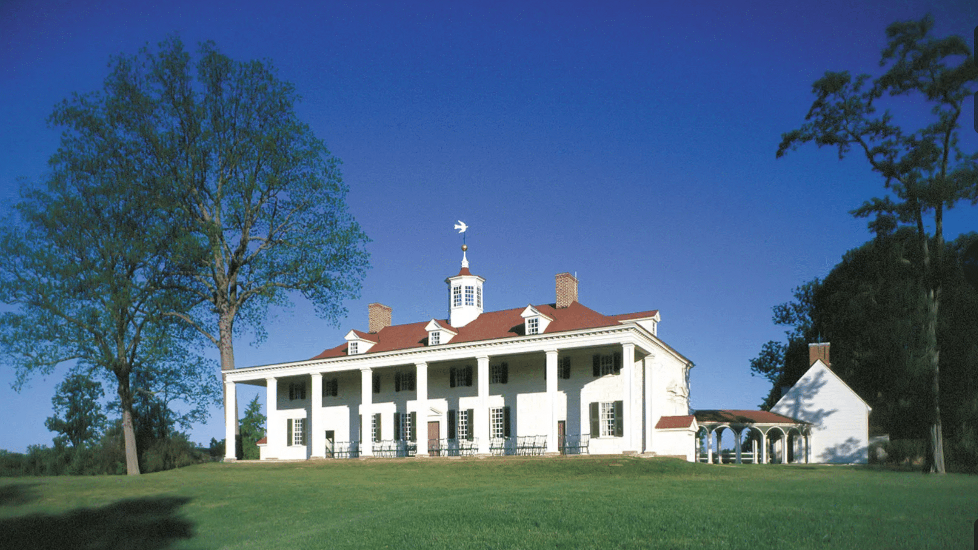 Large white mansion with a red roof and many pillars stands on a wide green field with tall trees and a clear blue sky