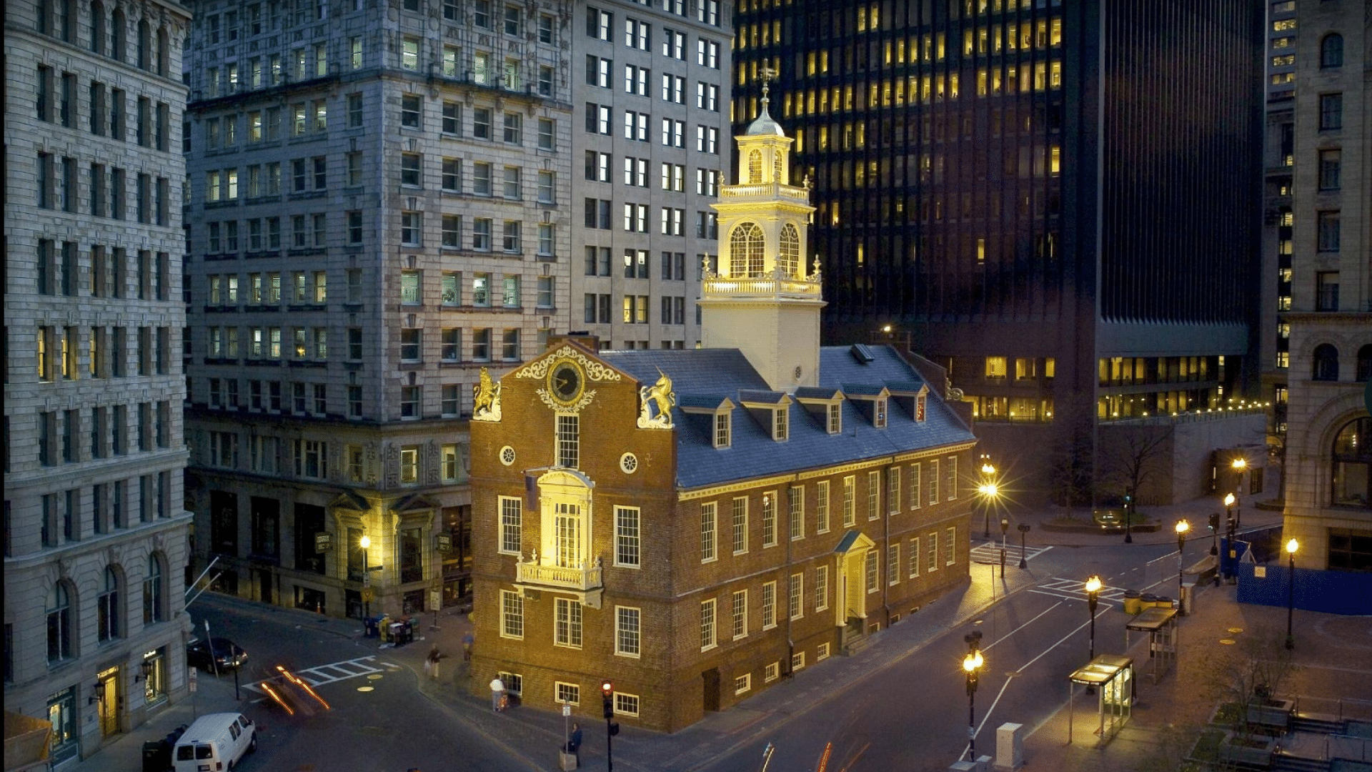 Old State House in Boston with a glowing white tower and gold statues is surrounded by tall city buildings at night time