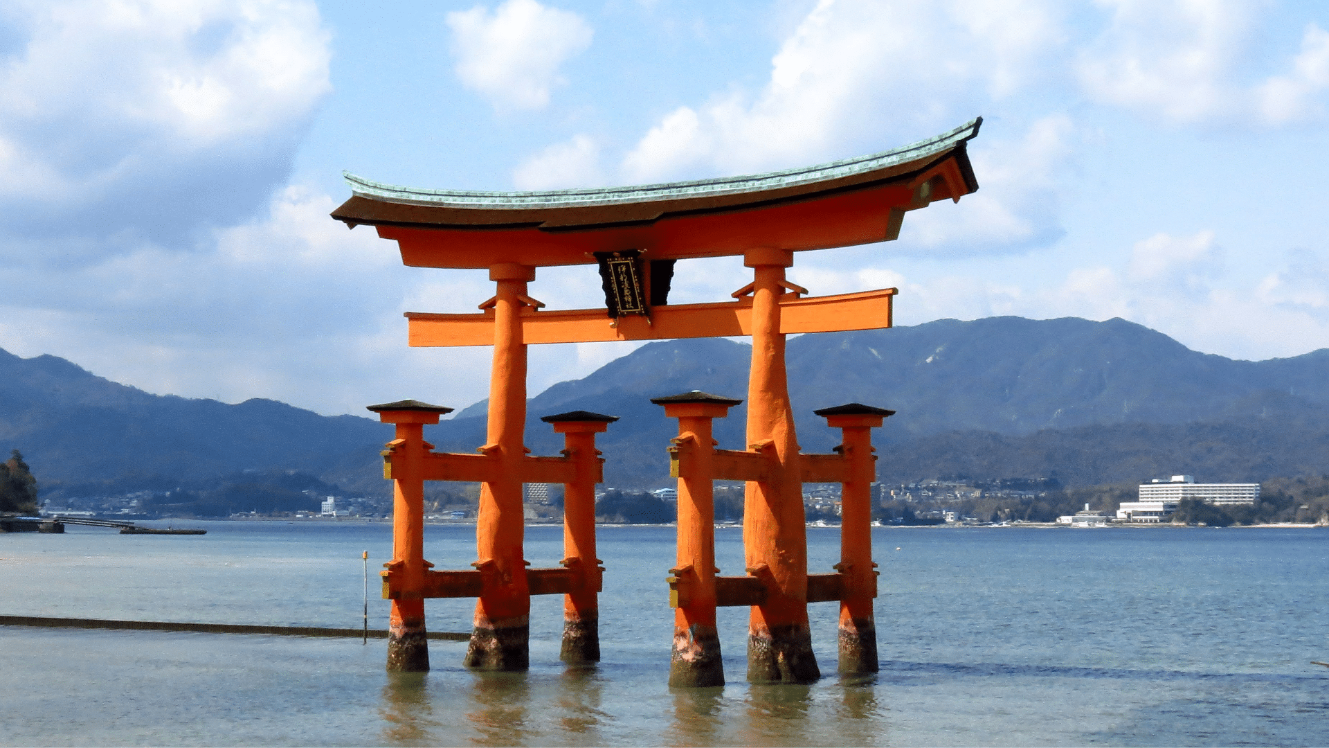 Orange Itsukushima torii gate stands in blue water with mountains behind and placed within a clear sunny sky
