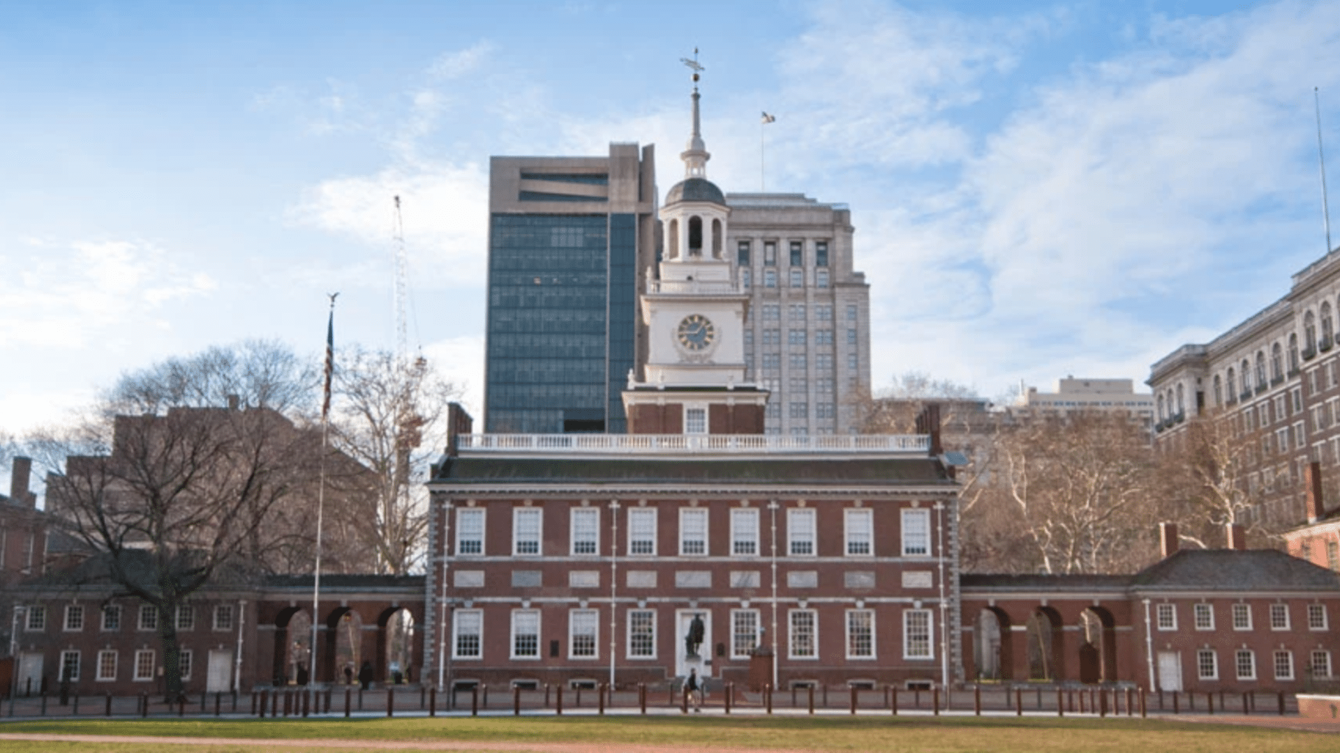 Red brick colonial building with a white clock tower and arched walkways under a blue sky with soft white clouds and trees