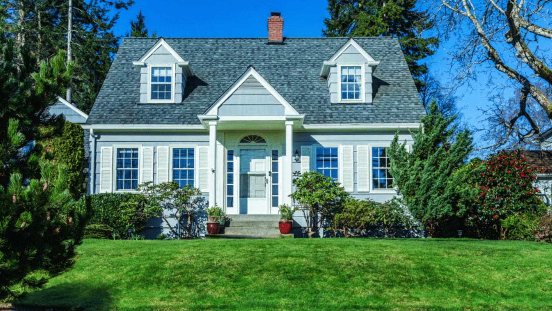 Small blue grey house with two dormer windows and white shutters on a sunny green lawn with tall trees and a blue sky