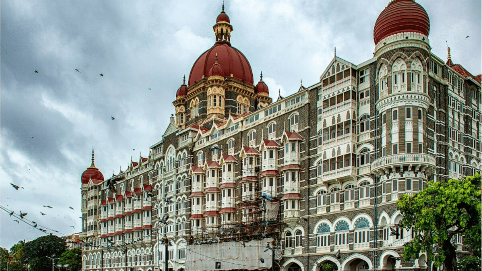 Taj Mahal Palace Hotel in Mumbai with red domes and many balconies under a cloudy sky with birds flying near green trees