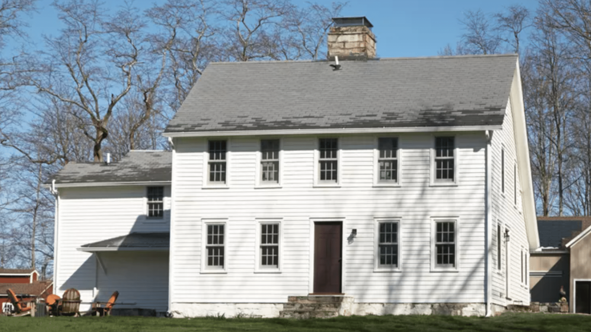 Two story white farmhouse with a grey roof and a stone chimney stands in front of bare winter trees under a clear blue sky