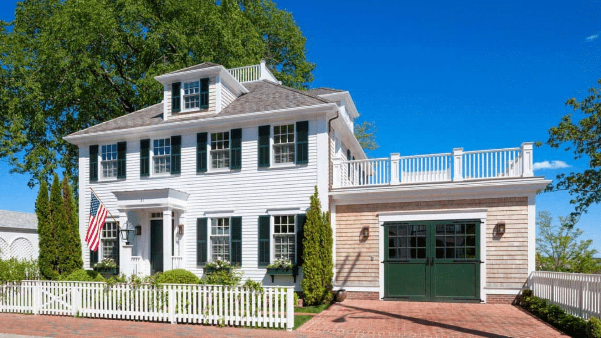 White colonial house with dark green shutters an American flag and a white picket fence under a clear blue summer sky
