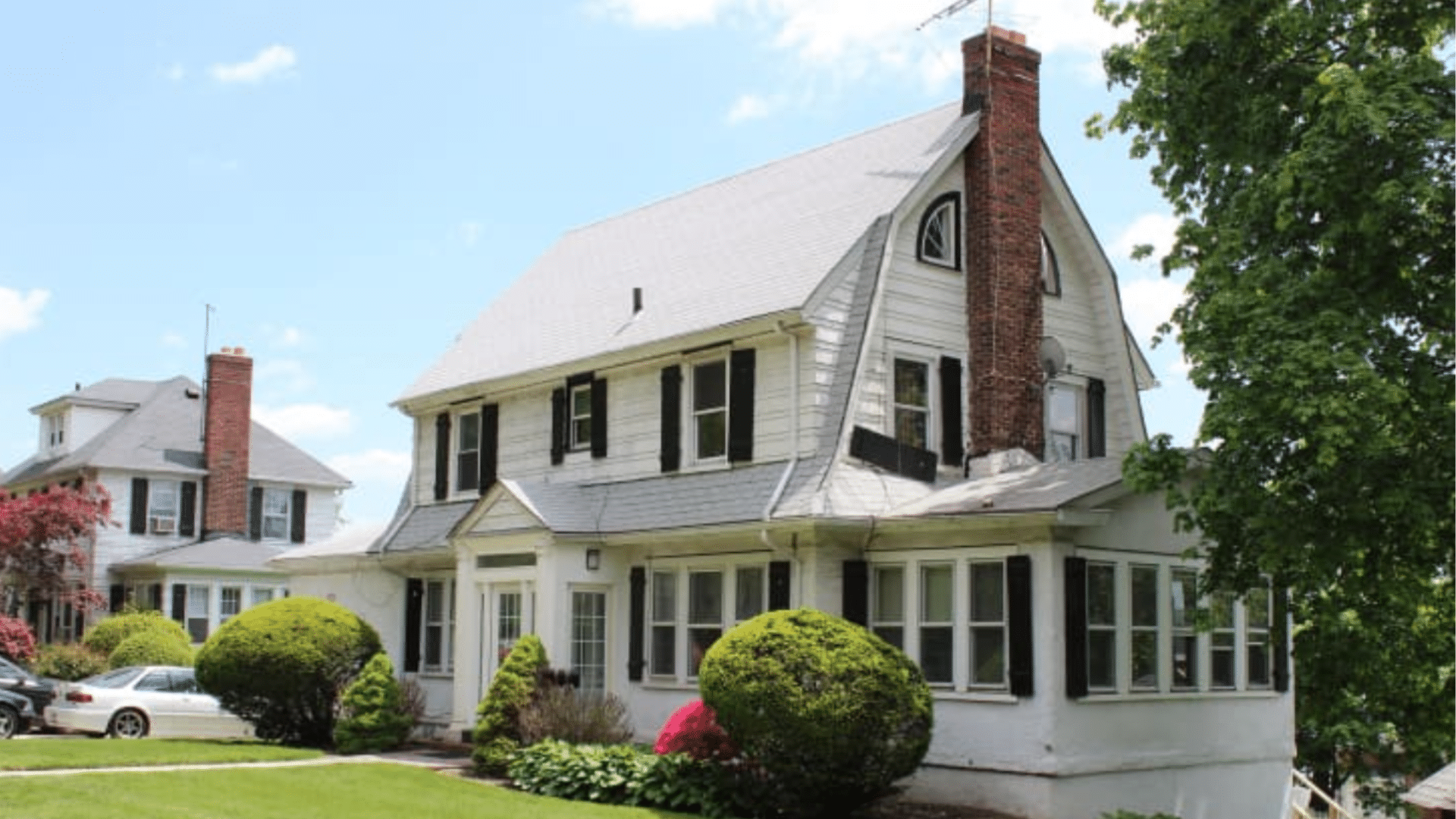White house with a brick chimney and black shutters has a gambrel roof and green bushes under a bright blue summer sky