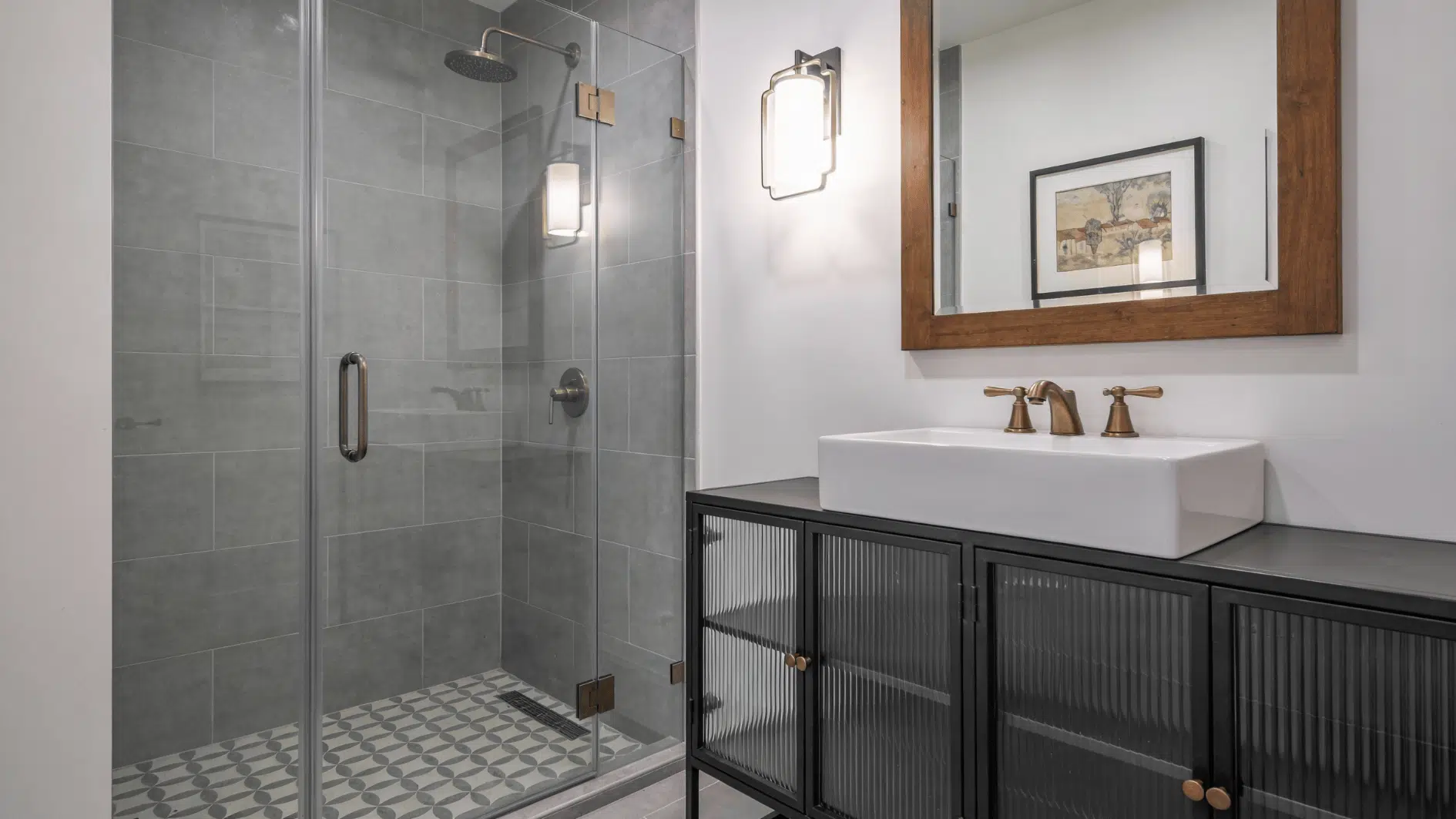 A frameless glass divider separates a gray tiled walk in shower from a modern vanity, keeping the small bathroom open and bright