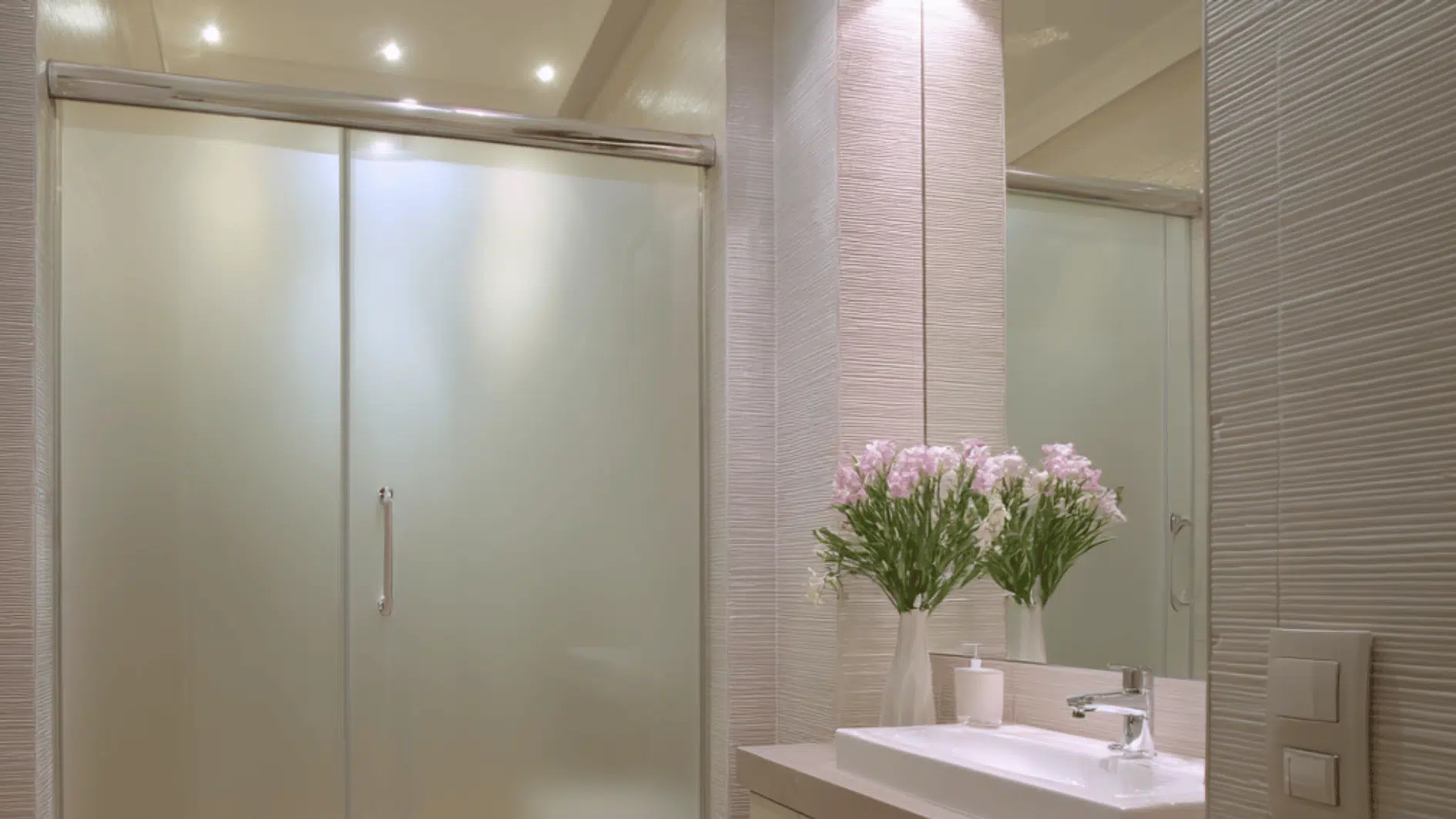 A modern bathroom with a walk in shower featuring frosted glass doors, soft lighting, and a sleek vanity with fresh flowers