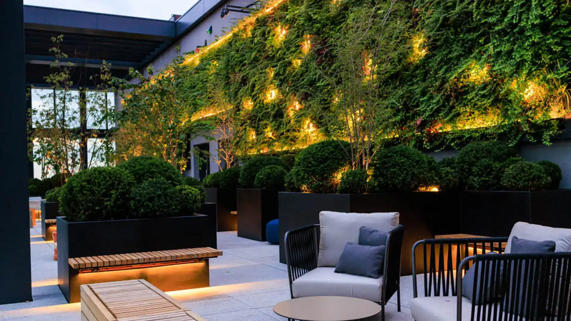 Rooftop patio featuring modern black planters with lush greenery, integrated wooden benches with warm underlighting, and an illuminated ivy wall at dusk