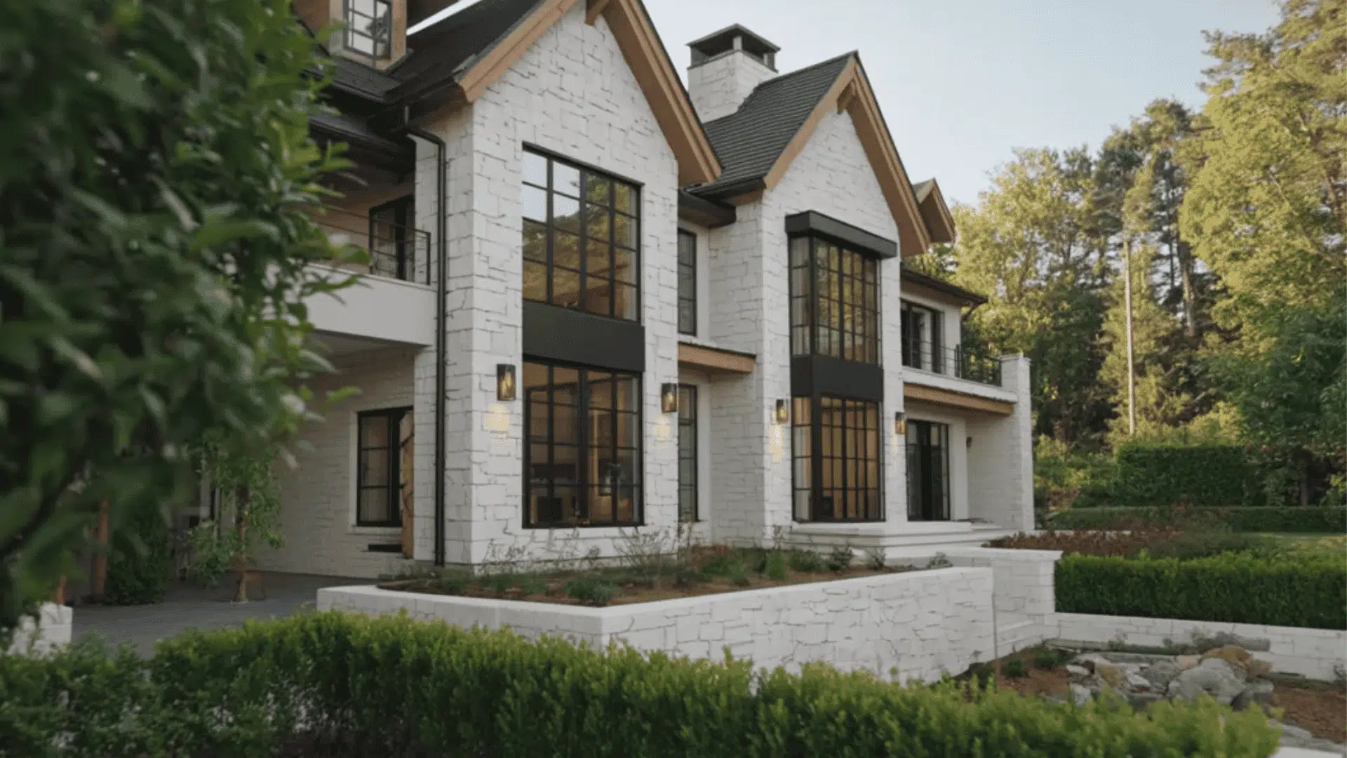 White brick house with large black windows, sloped roof, and greenery in a calm outdoor setting