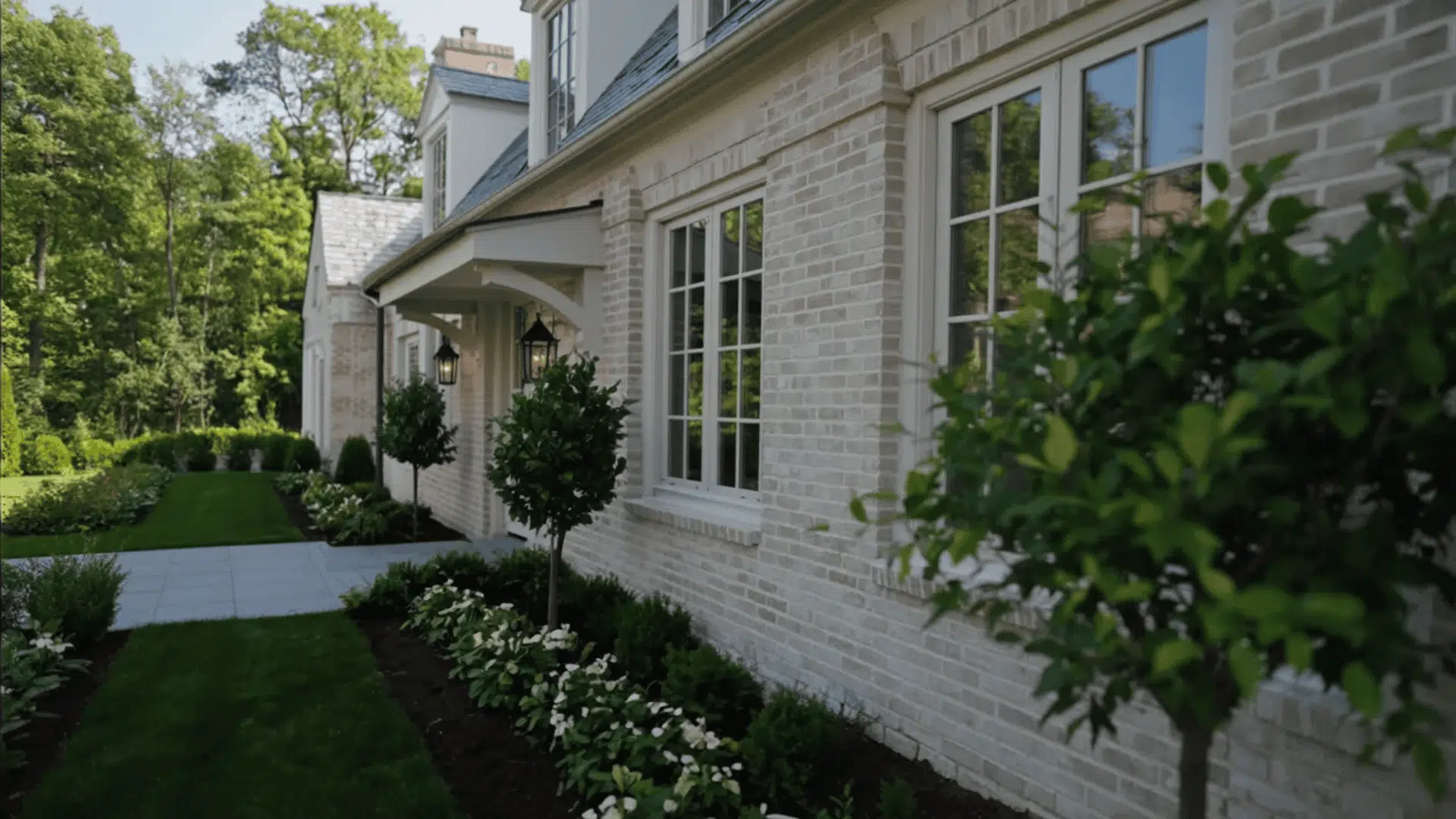 White brick house side view with windows, neat garden path, trimmed shrubs, and greenery along the wall
