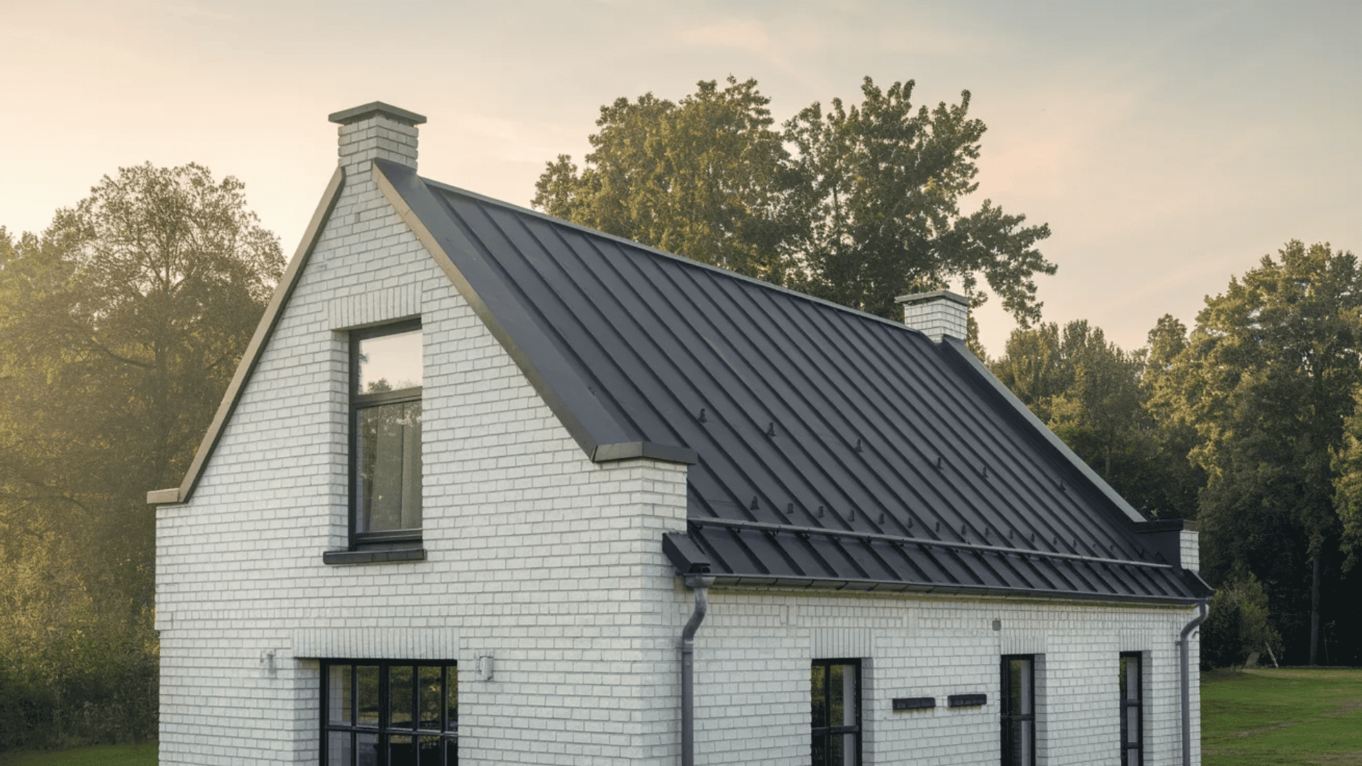 White house with steep black roof, tall window, and trees in soft evening light