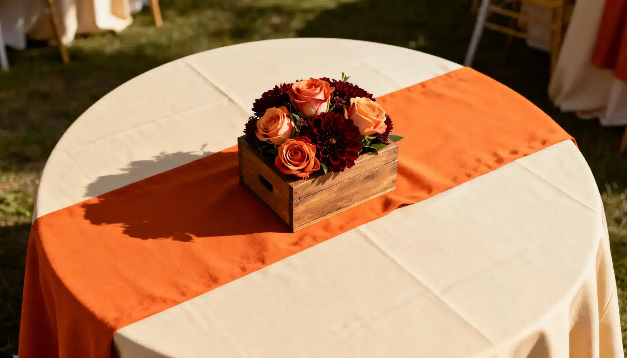 A wooden box with orange roses and dark red flowers sits on a round table, covered with a cream and orange tablecloth, bathed in warm sunlight