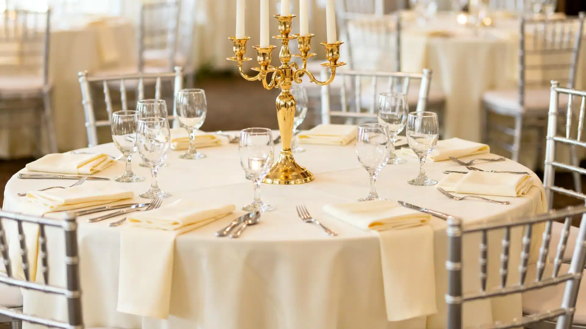 Banquet table set with a beige tablecloth, silver chairs, and a golden candelabra centerpiece with glasses and cutlery neatly arranged.