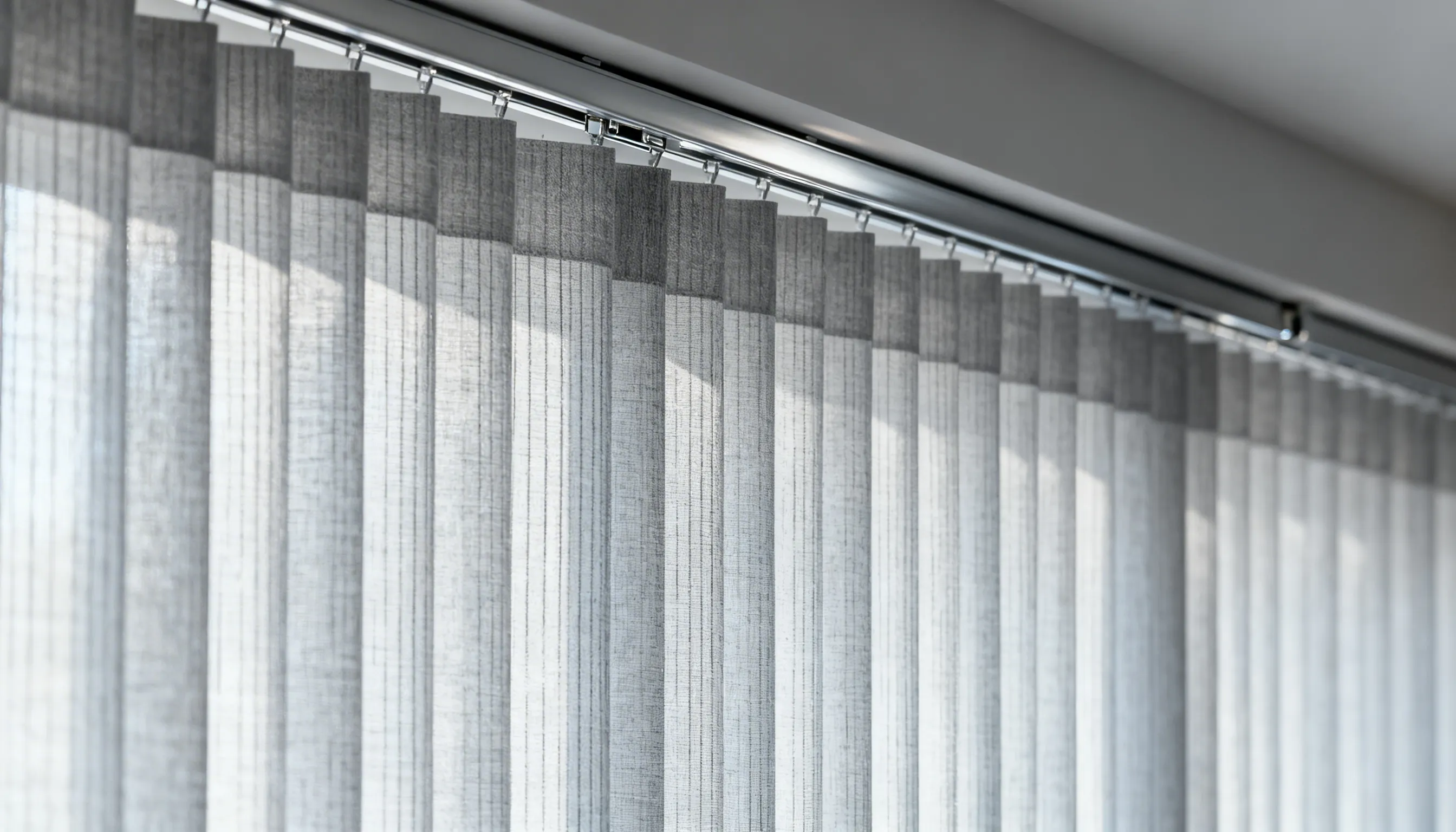 Close-up of light gray vertical blinds with fabric slats hanging from a metallic headrail against a bright window