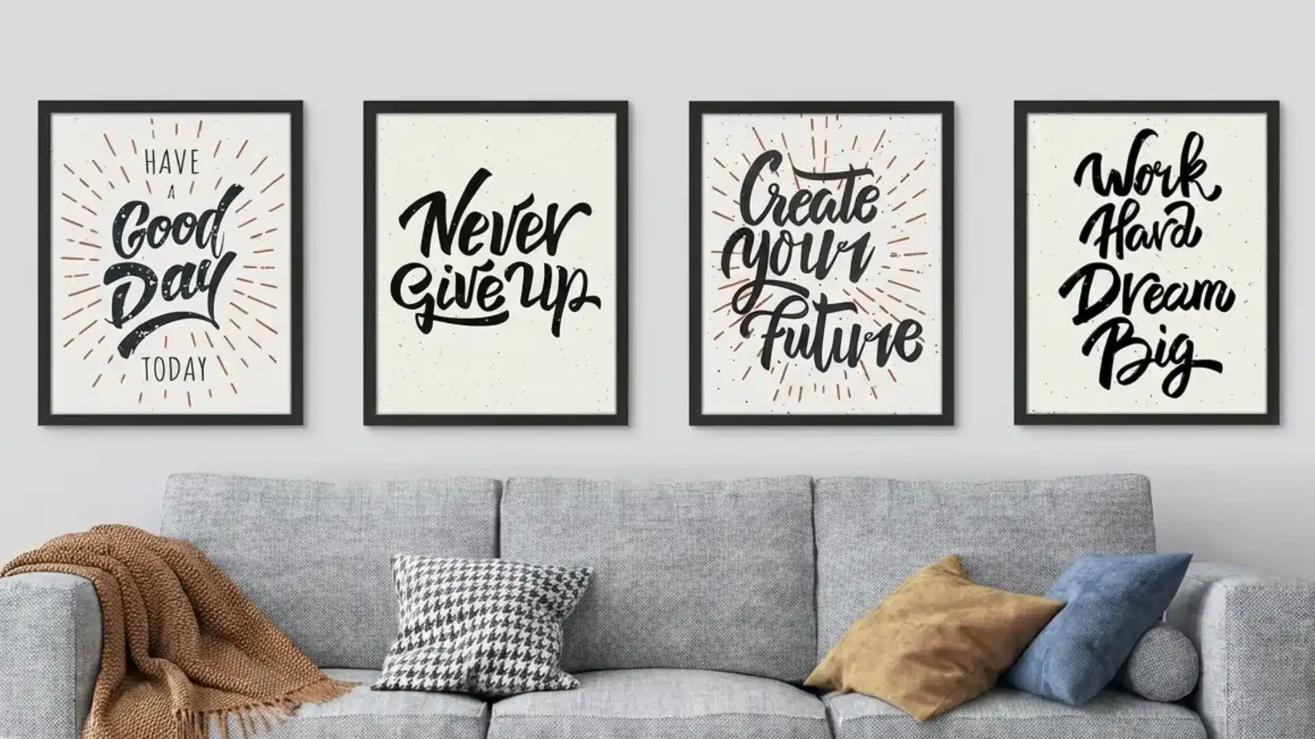 Four framed motivational posters of word art hang above a gray sofa adorned with throw pillows and a brown blanket
