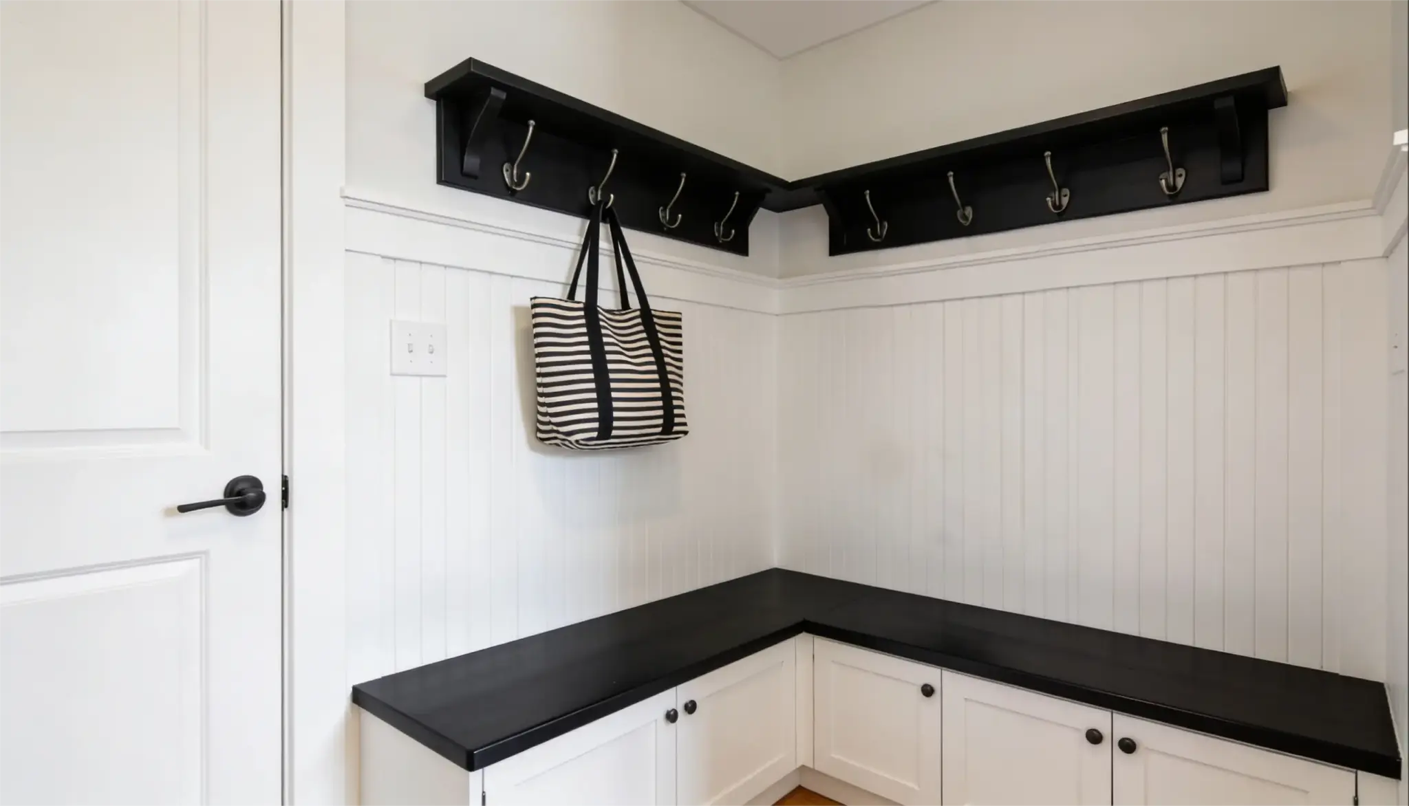 A modern mudroom corner featuring white beadboard walls, white lower cabinets with black knobs and a black wall-mounted coat rack with a striped tote bag hanging on one hook