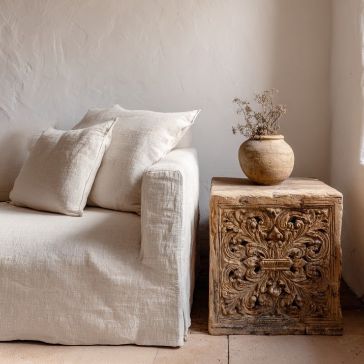 Neutral linen sofa beside carved wood side table, white plaster wall, modern Spanish interior style