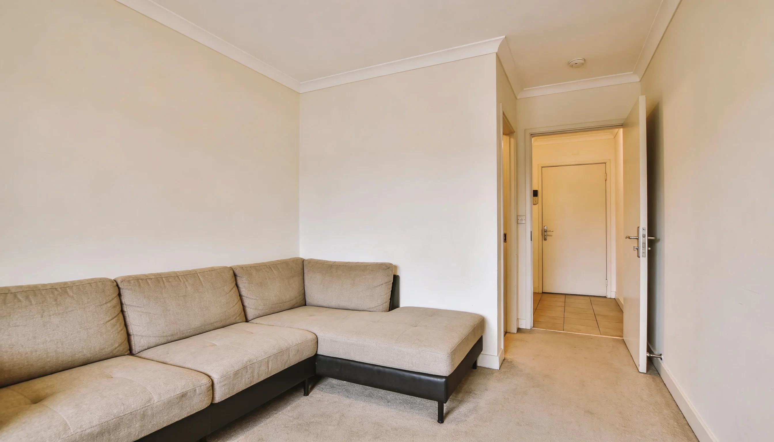 A beige sectional sofa sits against a light-colored wall next to a hallway leading to an open doorway with a light-colored interior door