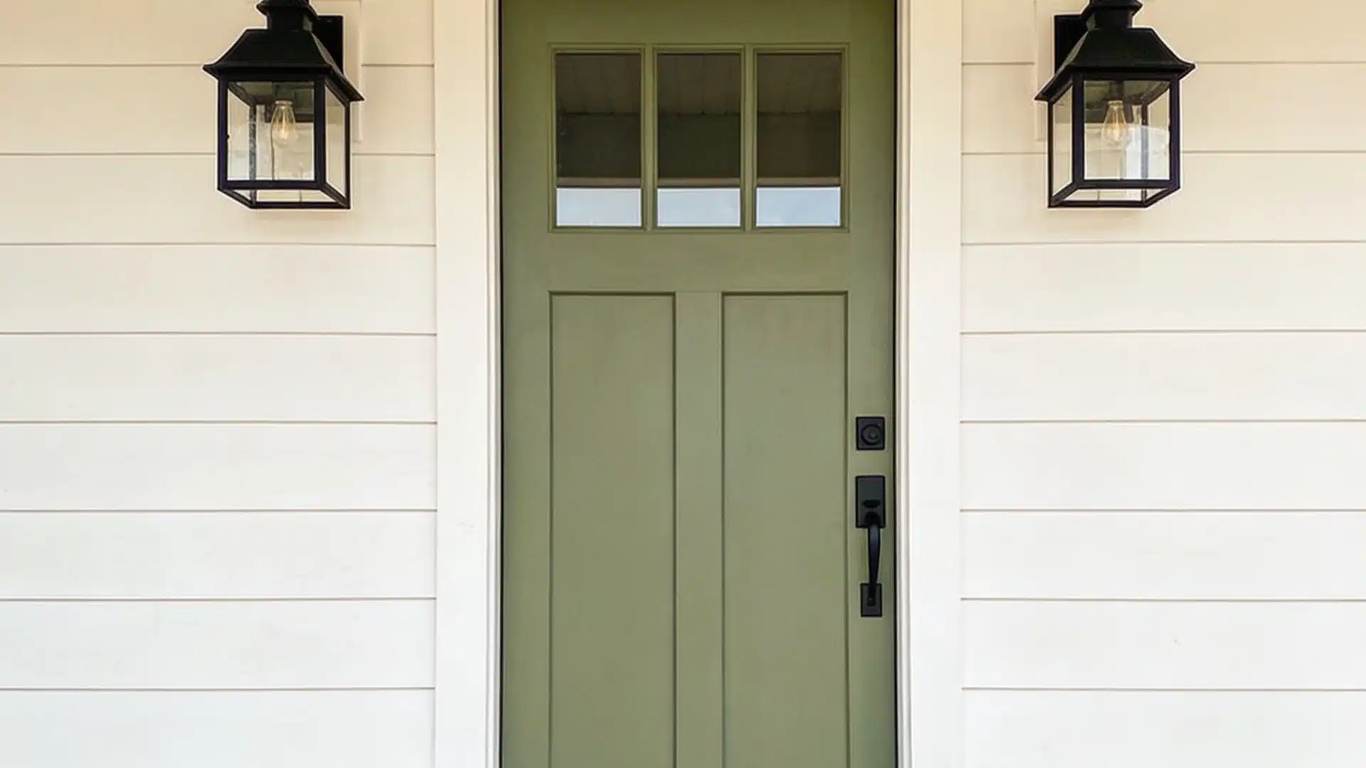 A front view of a sage green entry door with four top window panes, flanked by black lantern sconces on white shiplap siding, above a natural wood porch floor.