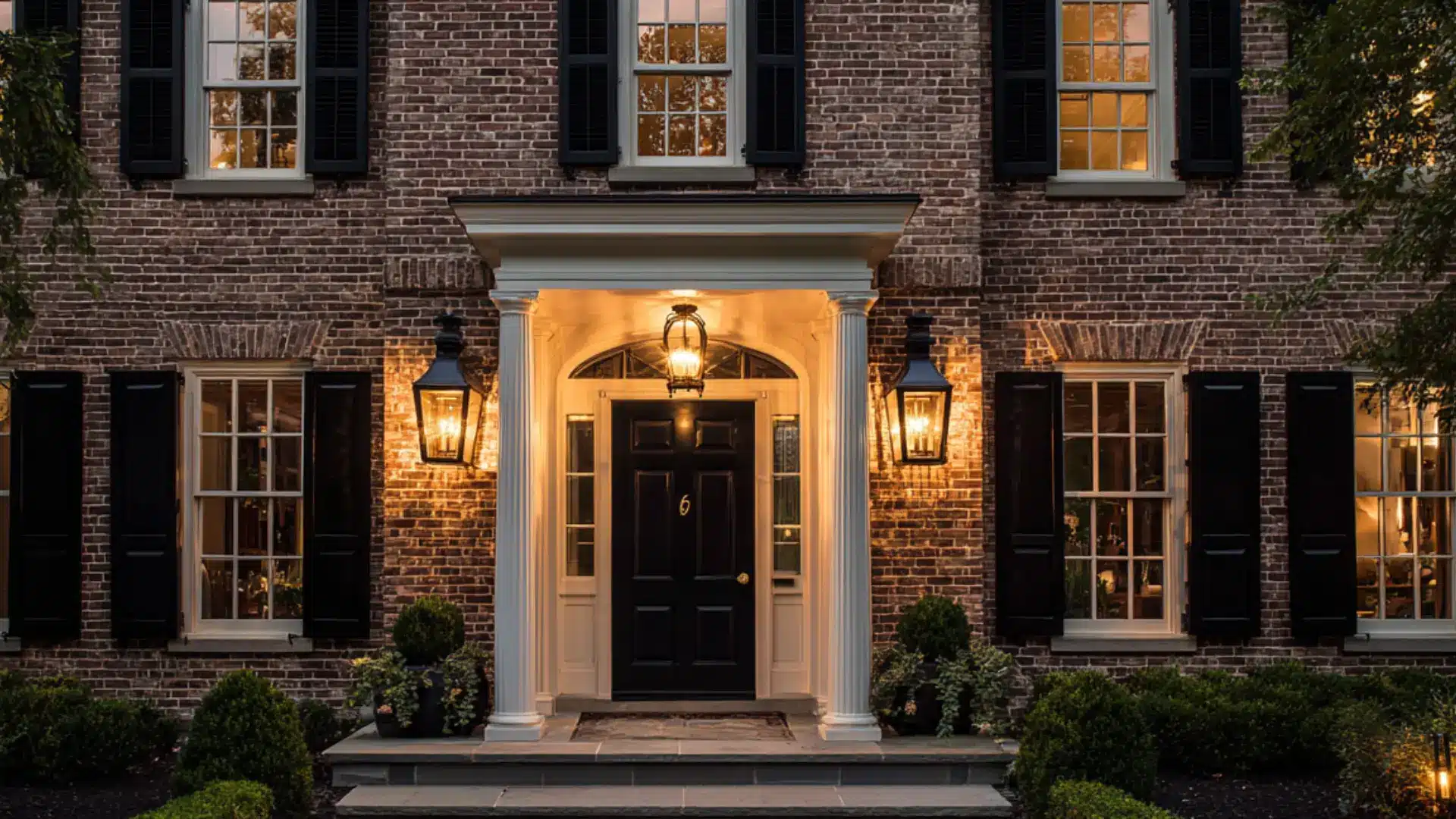Brick house at dusk featuring black shutters a white portico with columns warm lantern light and a stone garden walkway