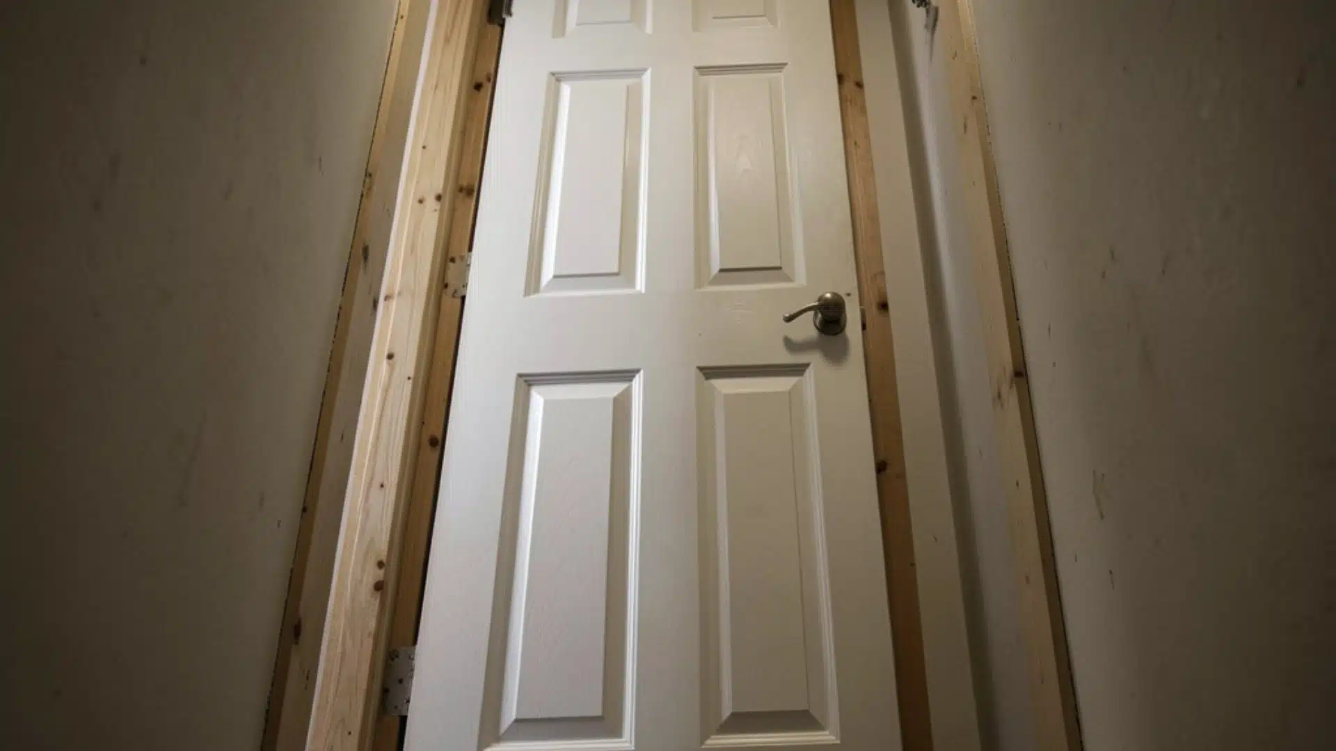 Low-angle view looking up a narrow hallway toward an open, six-panel white door framed by raw wooden studs.