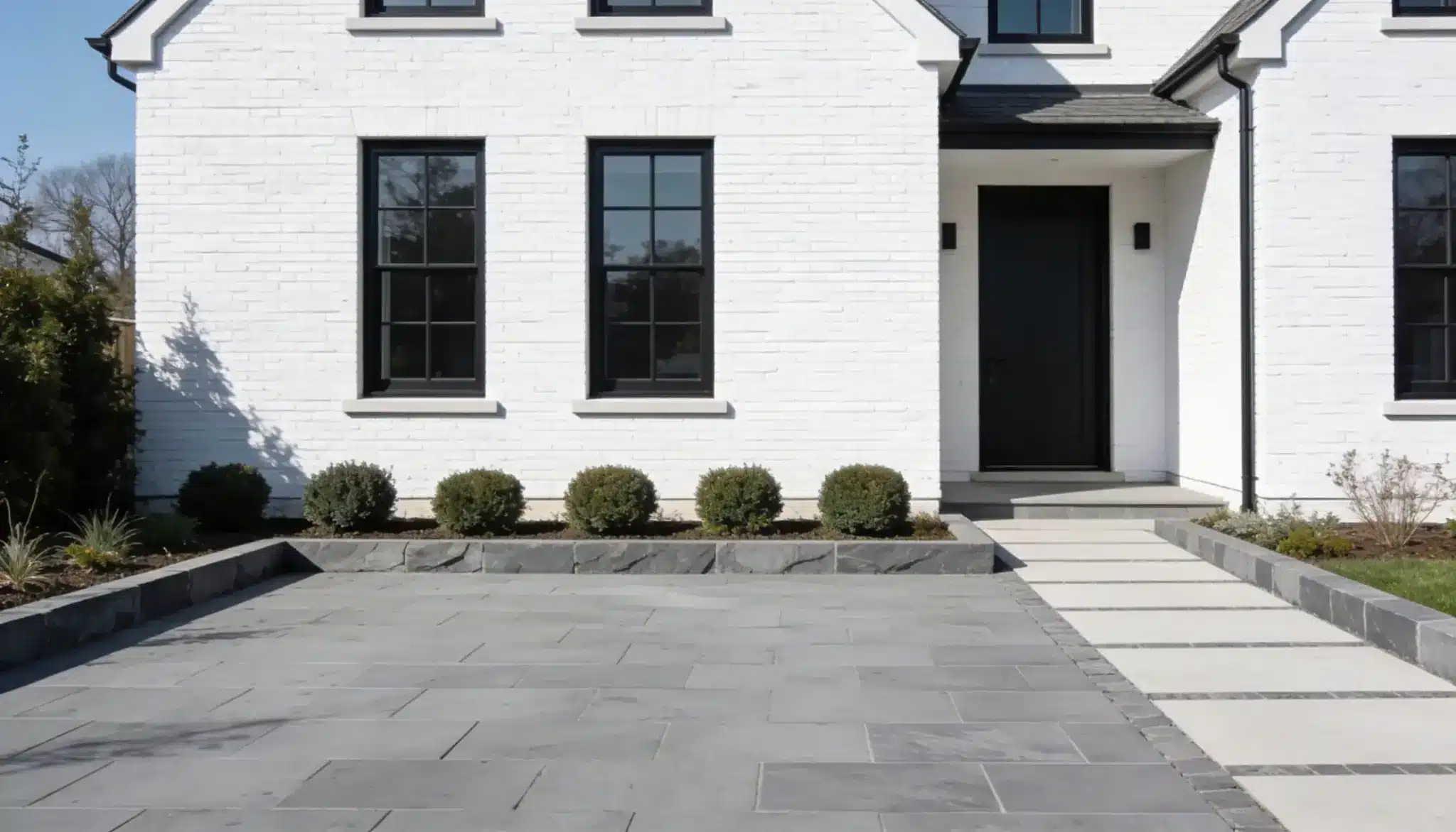 Modern white brick house exterior featuring black trim windows and door, contrasting with a grey stone driveway and walkway bordered by manicured green shrubs