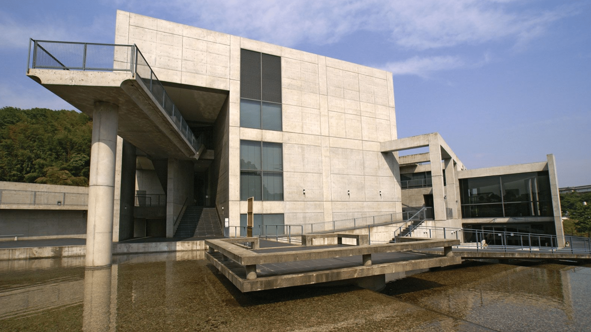 Modern grey concrete building with sharp geometric lines and walkways reflecting in a still pool of water under a blue sky