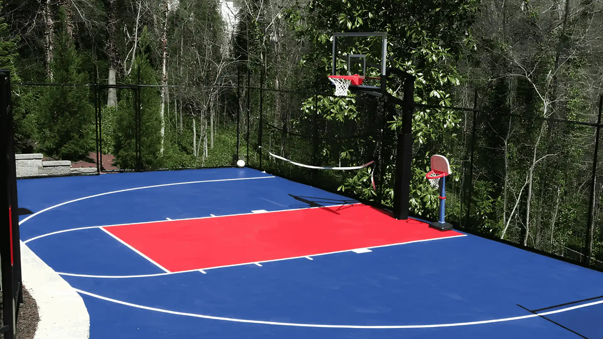 Blue outdoor basketball court with bright red key area and hoop beside wooded fencing