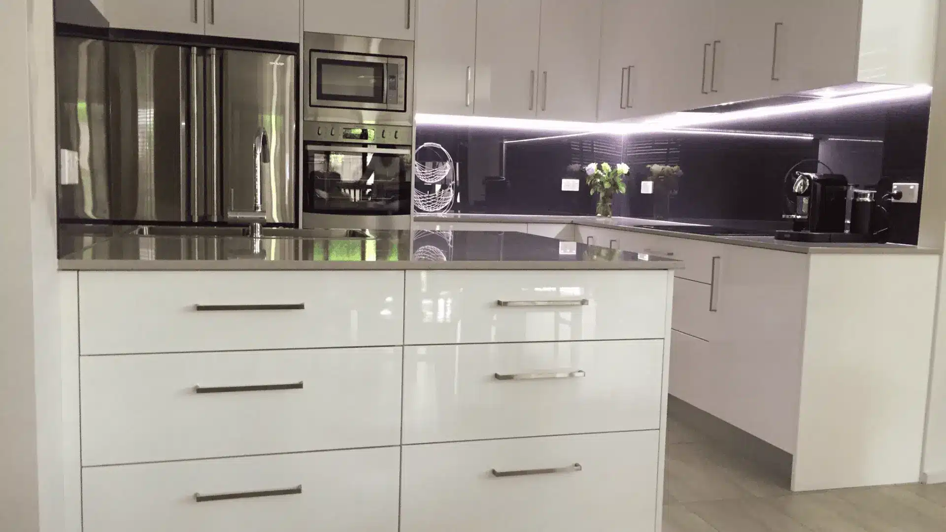 Modern white gloss kitchen with an island, silver handles, and a dark illuminated backsplash