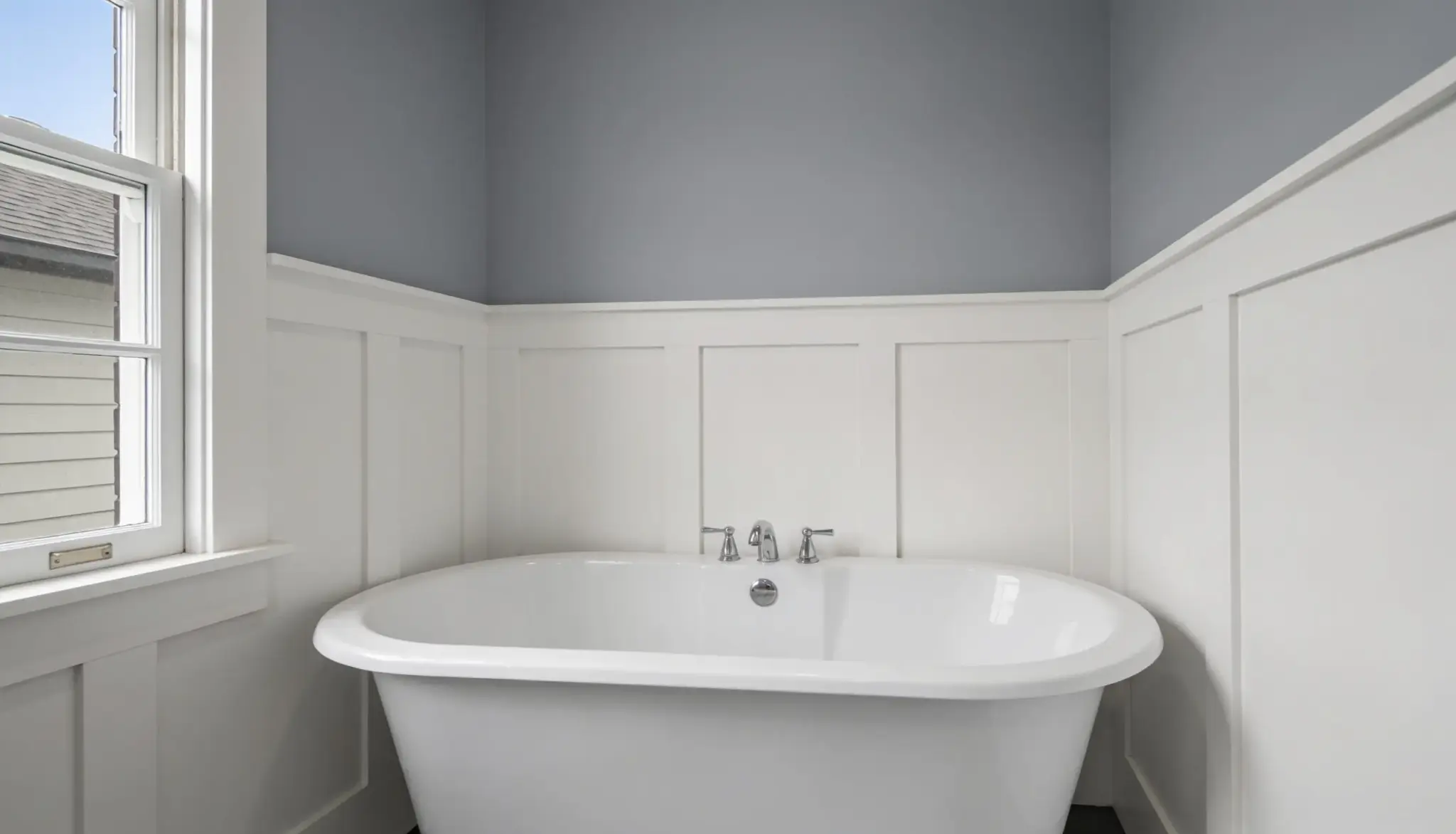 Freestanding white bathtub with chrome fixtures against a gray wall and white wainscoting, with a window to the left.