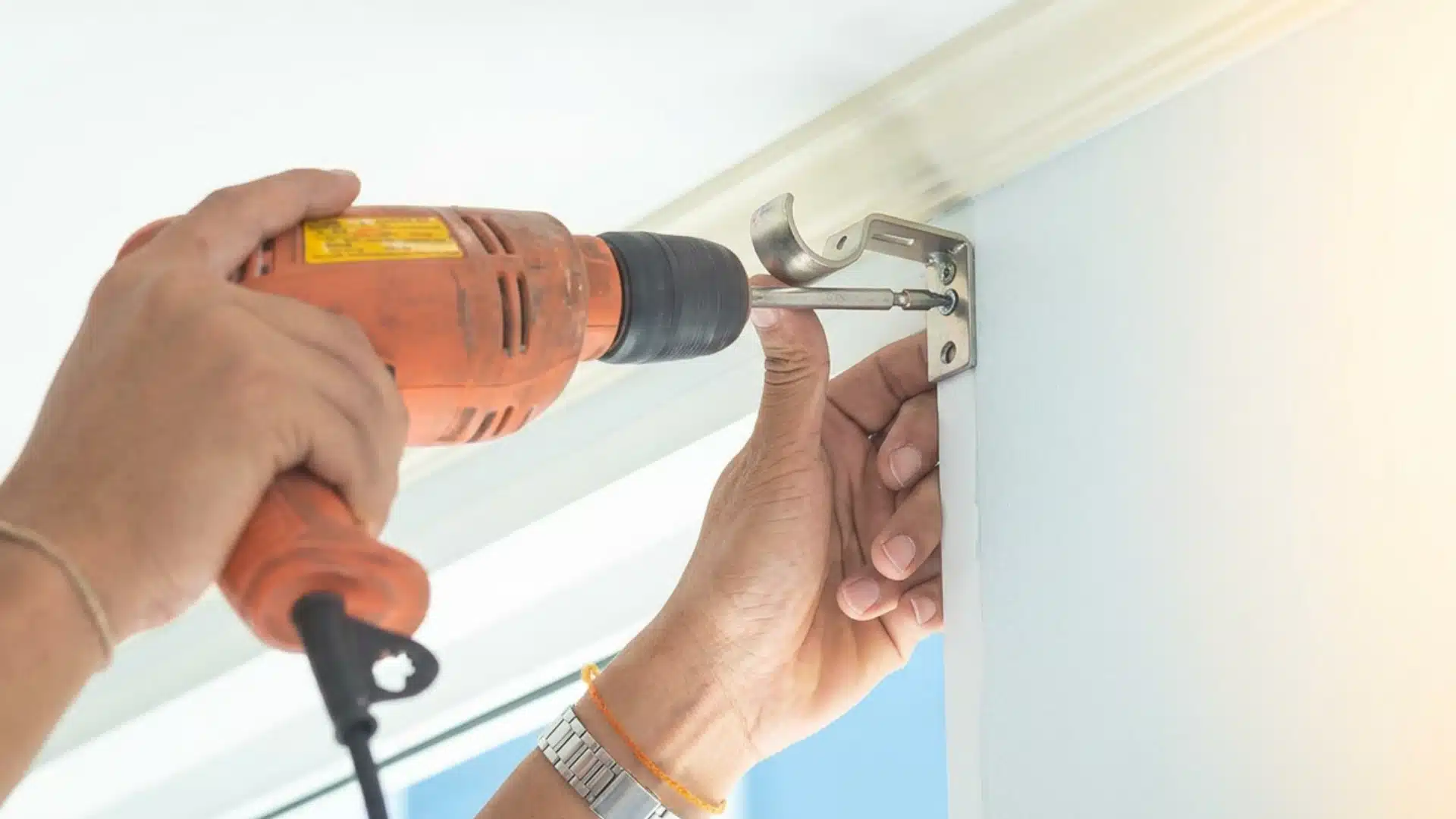 Hands use an orange power drill to screw a metal curtain rod bracket onto a white wall.