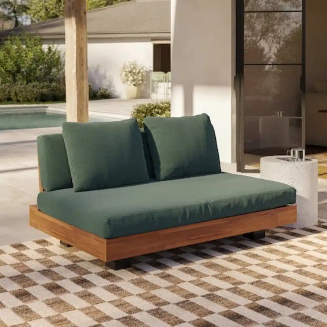Modern outdoor loveseat with deep green cushions, a warm wood frame, and a patterned rug beside a pool on a sunny patio.