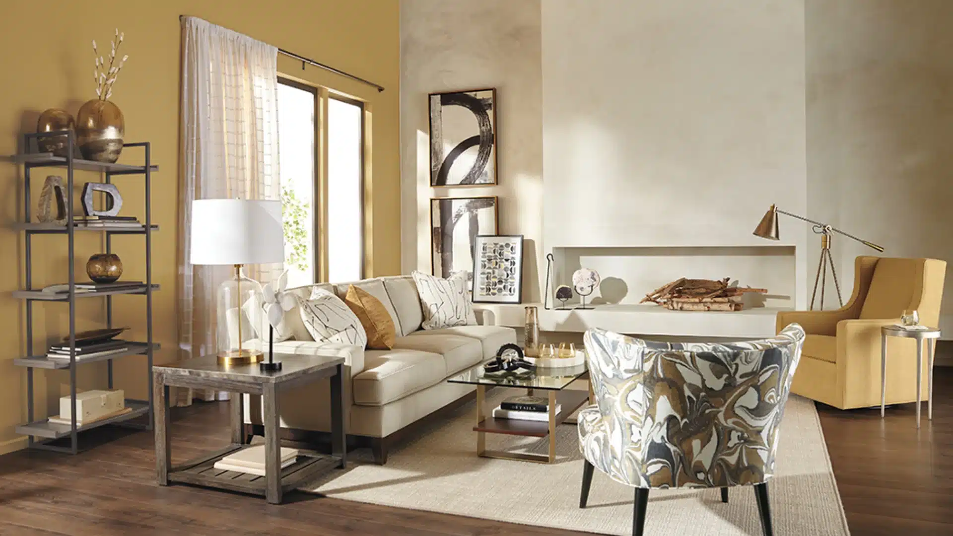 Warm modern living room with a cream sofa, patterned chair, mustard armchair, open shelving, abstract art, and wood flooring.