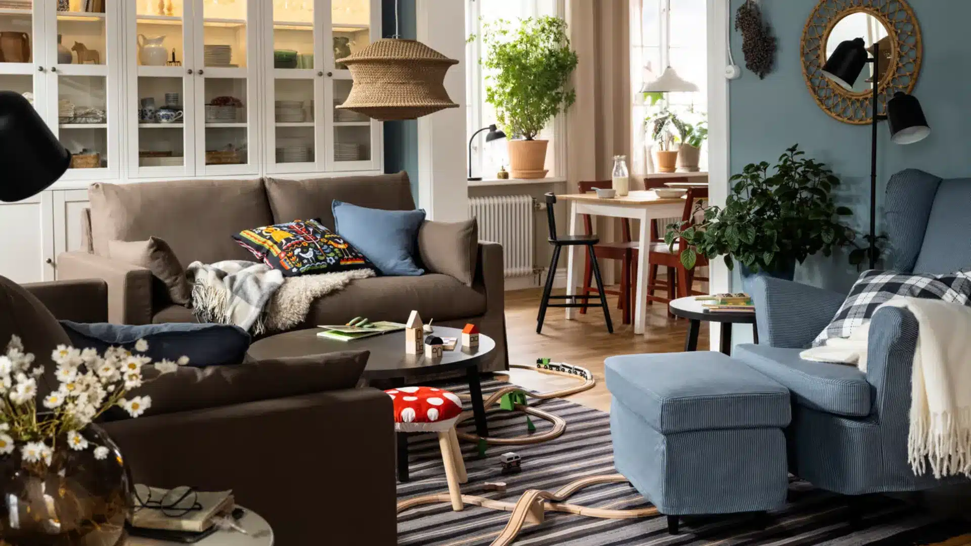 Cozy family living room with brown sofas, a blue armchair and ottoman, indoor plants, dining area, and children’s toys.