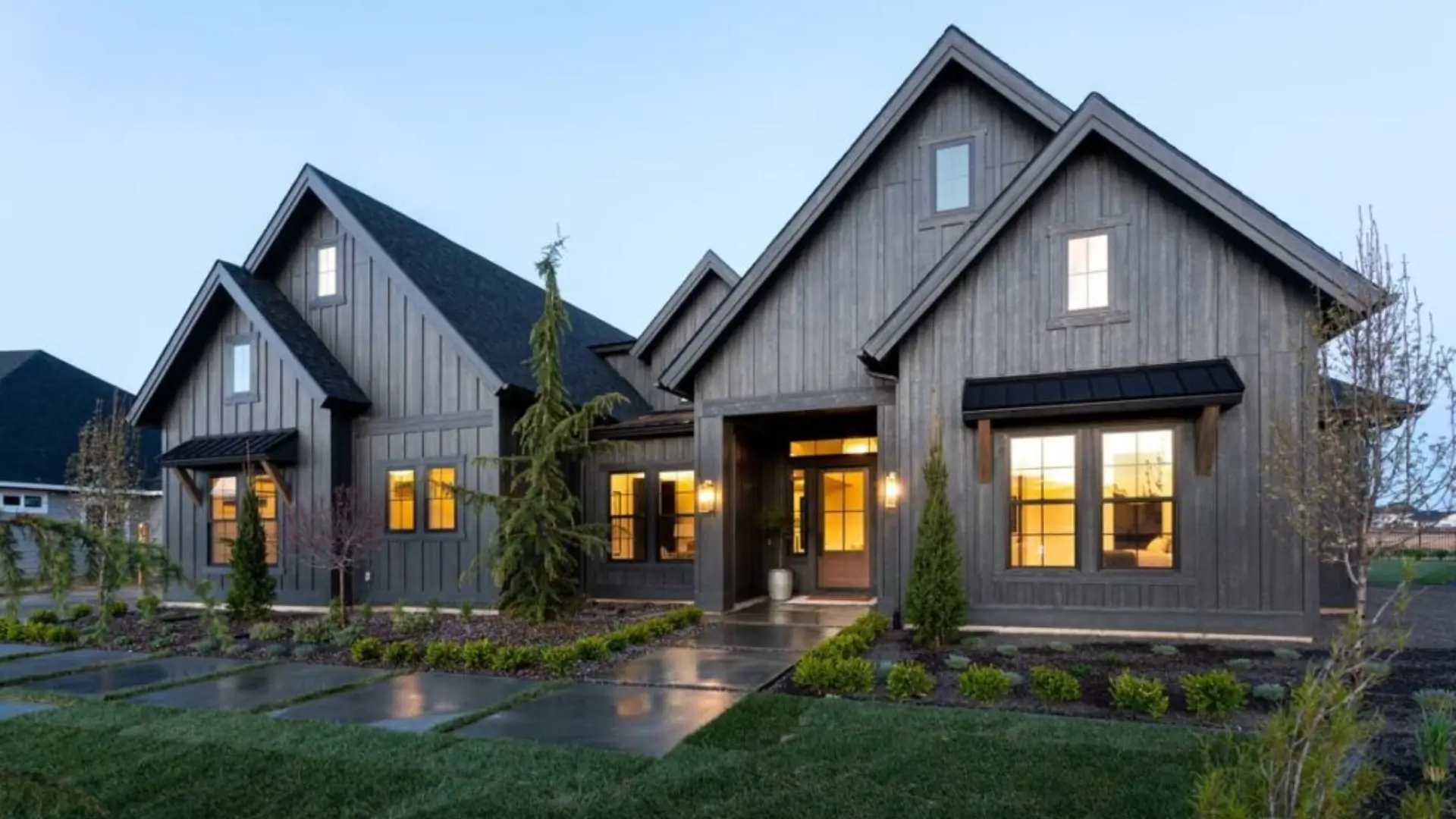 Modern gray-clad suburban house with peaked roofs, glowing windows, landscaped gardens, and wet walkways at dusk.