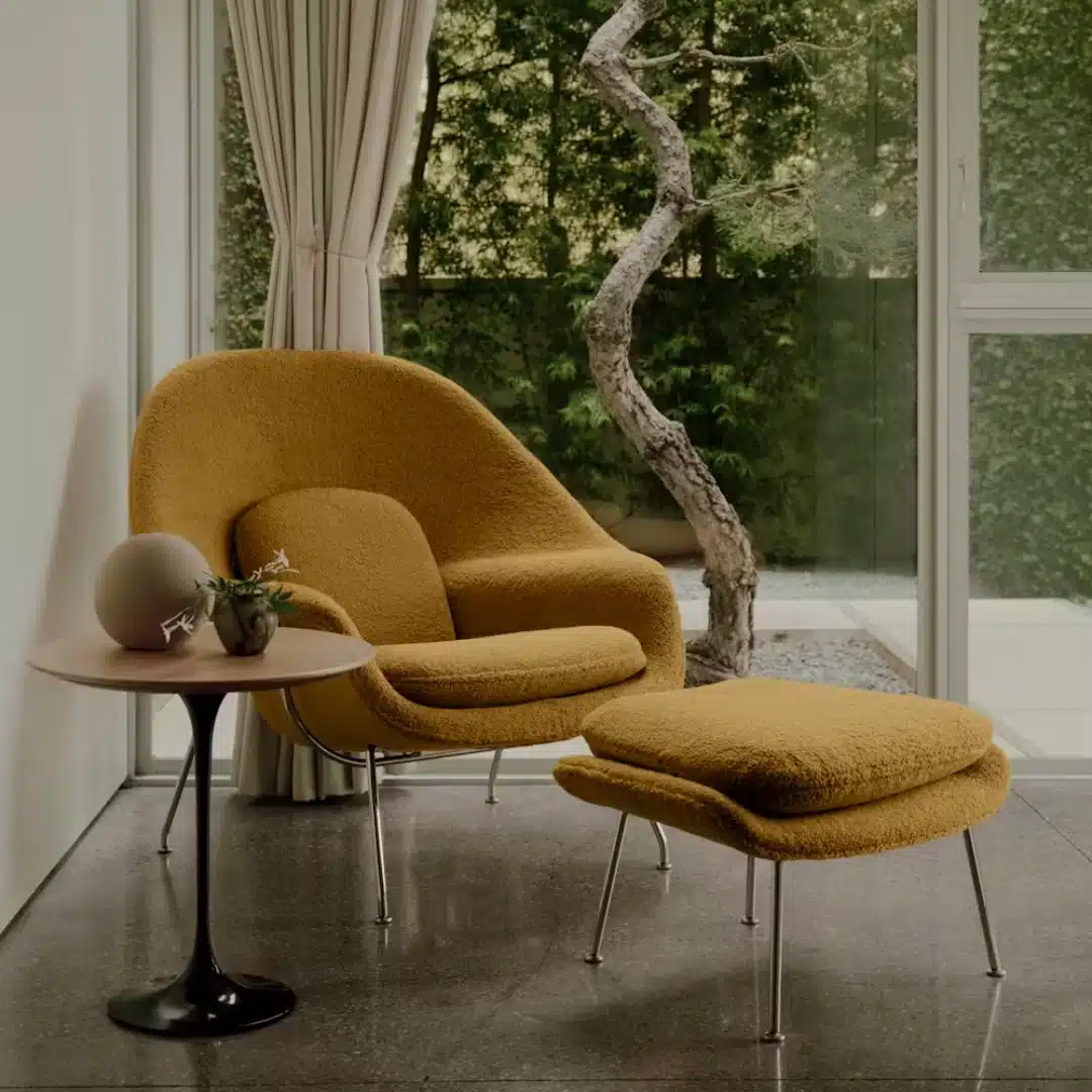 Mustard boucle lounge chair with matching ottoman and side table beside floor-to-ceiling windows overlooking a leafy garden.