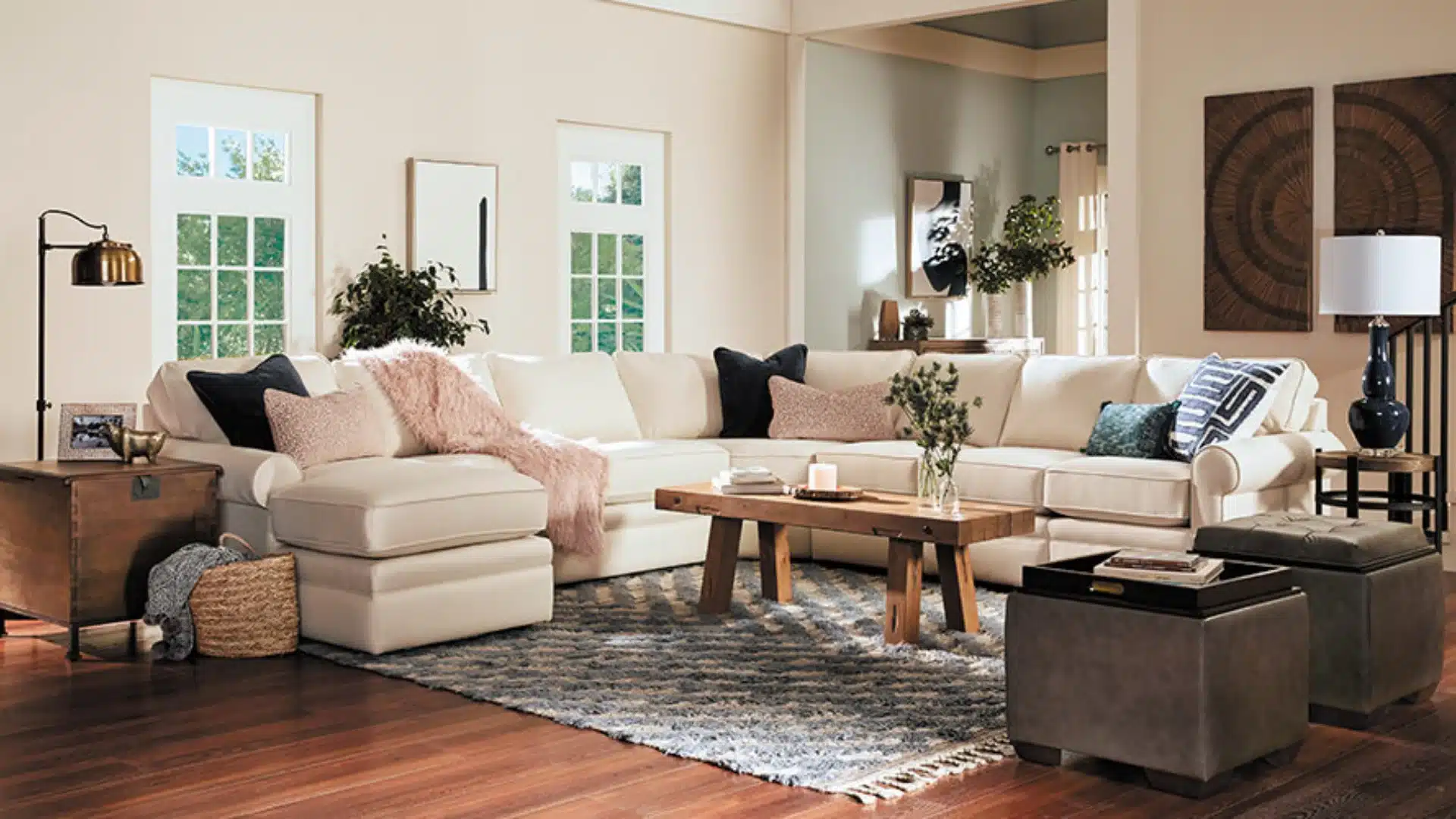 Bright living room with a large cream sectional sofa, wood coffee table, patterned rug, dark ottomans, lamps, and wall art.