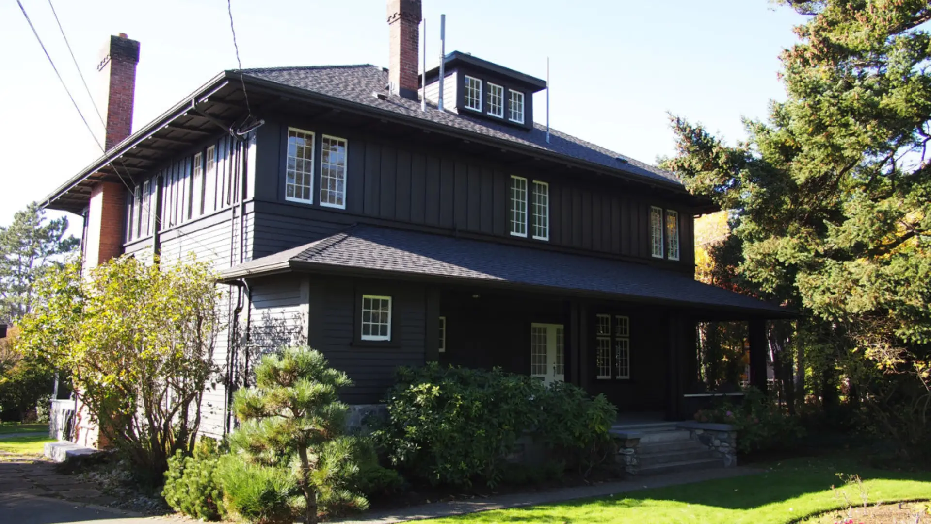 Dark gray two-story Craftsman-style house with white-trimmed windows, brick chimneys, a covered porch, and landscaped greenery.