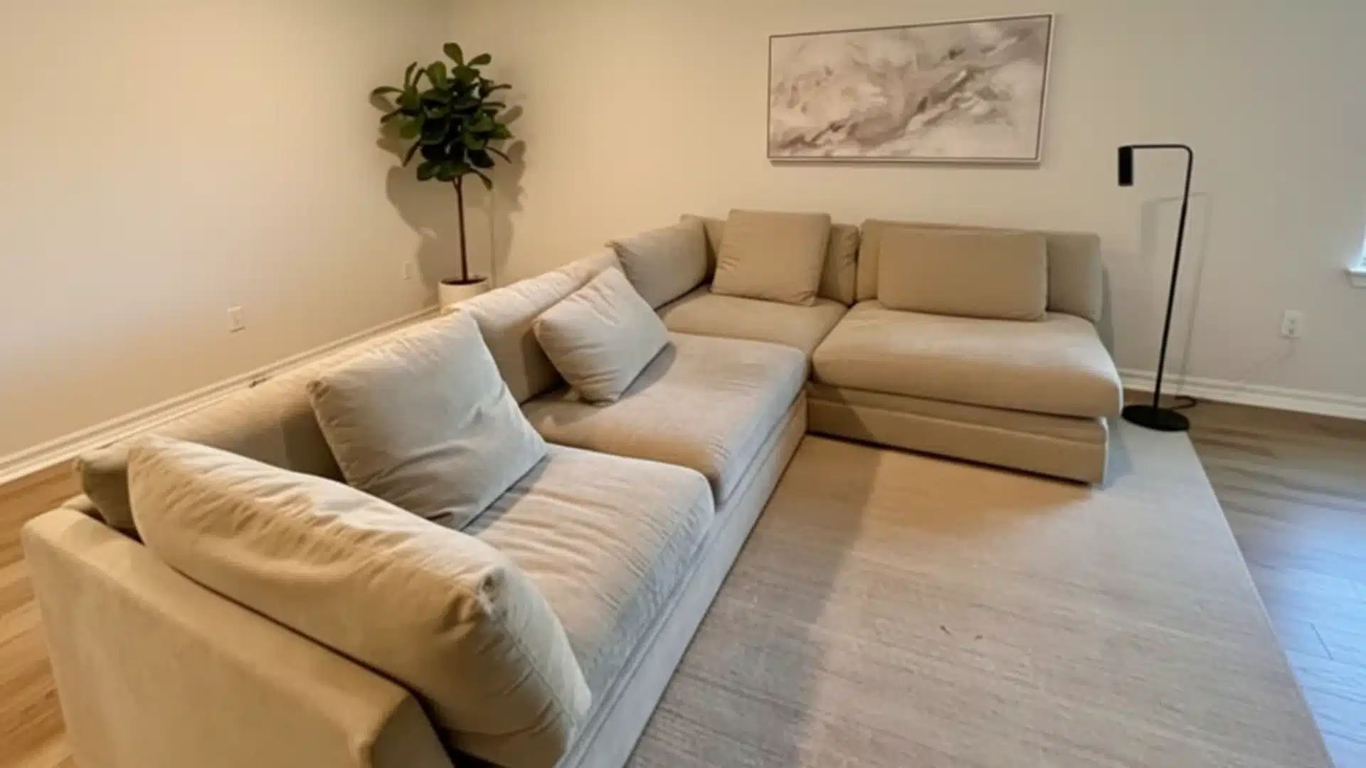 Beige sectional sofa with cushions on a light rug in a modern living room with a potted plant, abstract wall art, and black floor lamp.