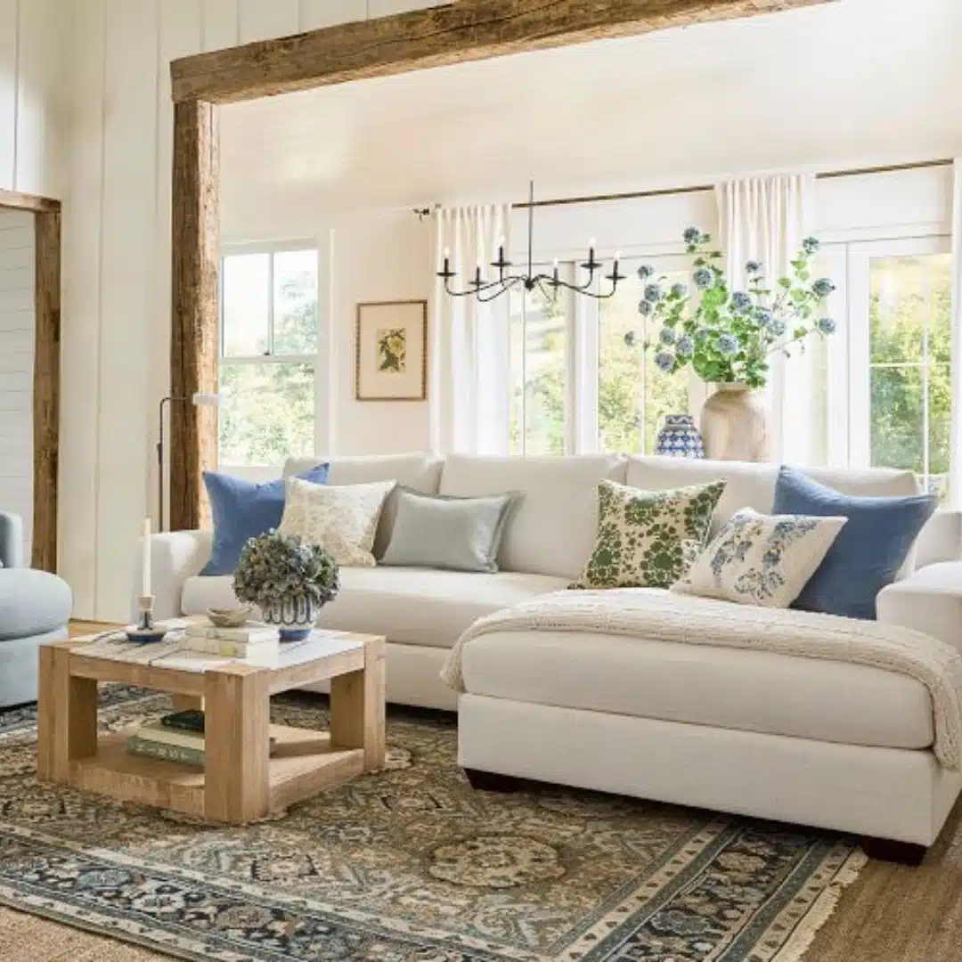 Bright living room with a cream sectional sofa, blue and patterned pillows, wood coffee table, chandelier, and large sunlit windows.