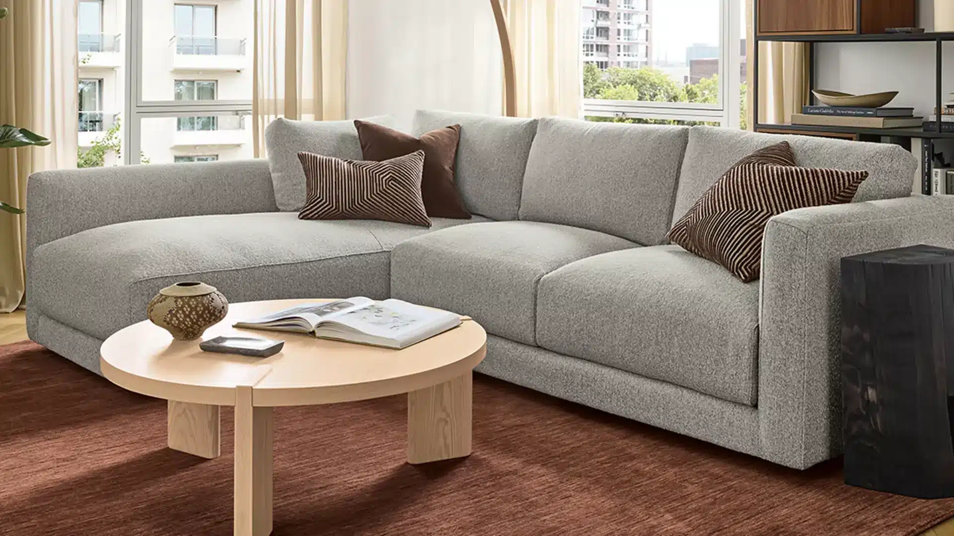 Light gray sectional sofa with brown pillows in a bright modern living room, paired with a round wood coffee table and rust rug.
