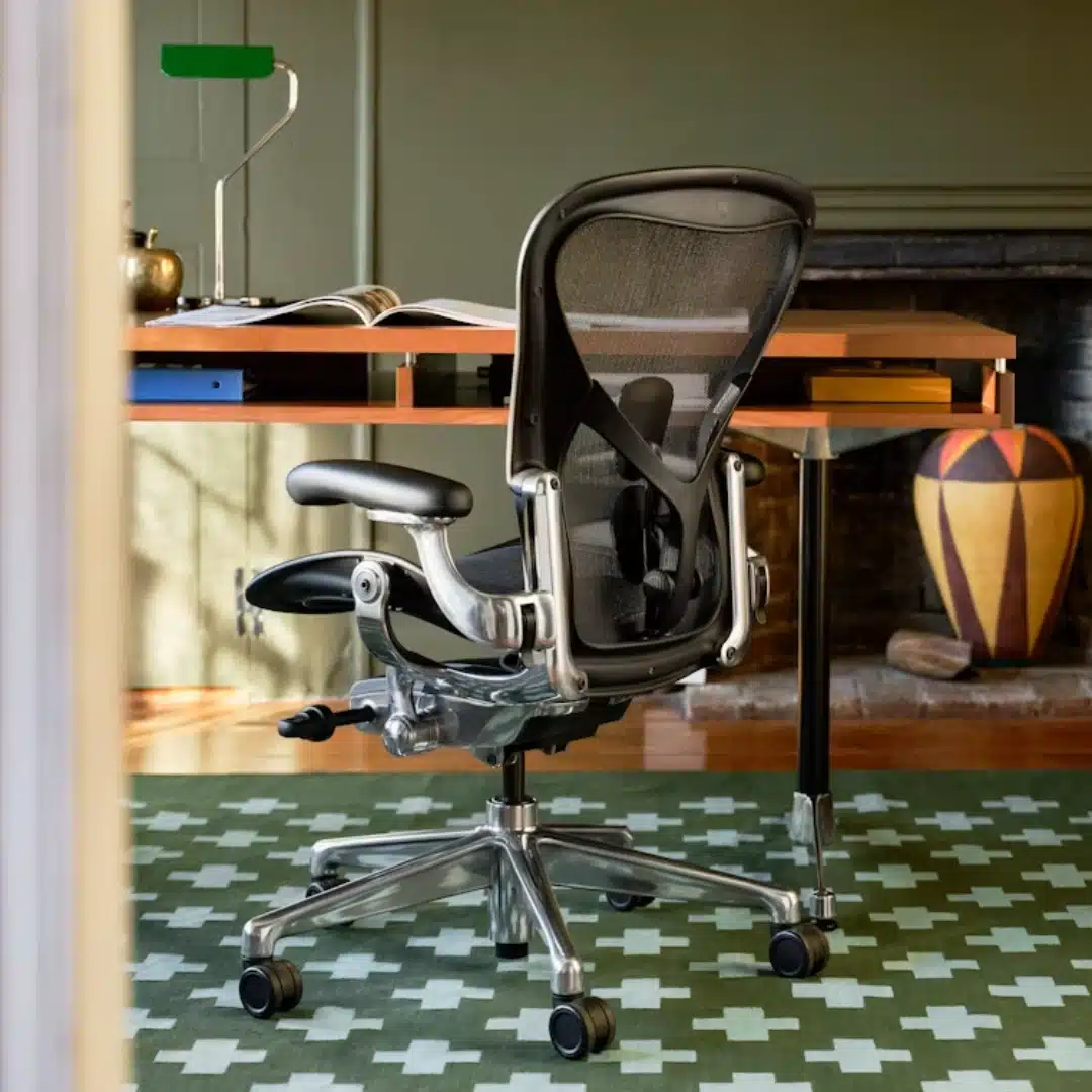 Ergonomic black mesh office chair with polished metal frame at a wood desk, set on a green patterned rug in a home office.