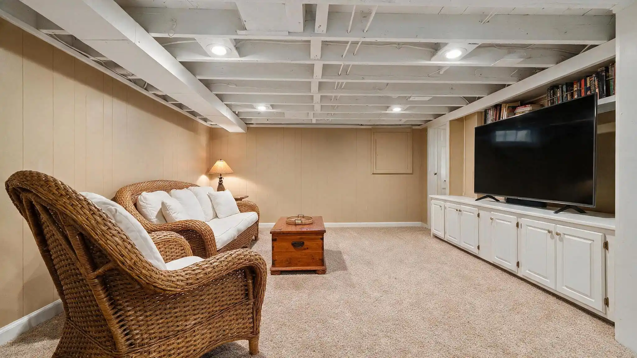 Finished basement living room with white exposed ceiling joists wicker seating television and soft neutral carpet