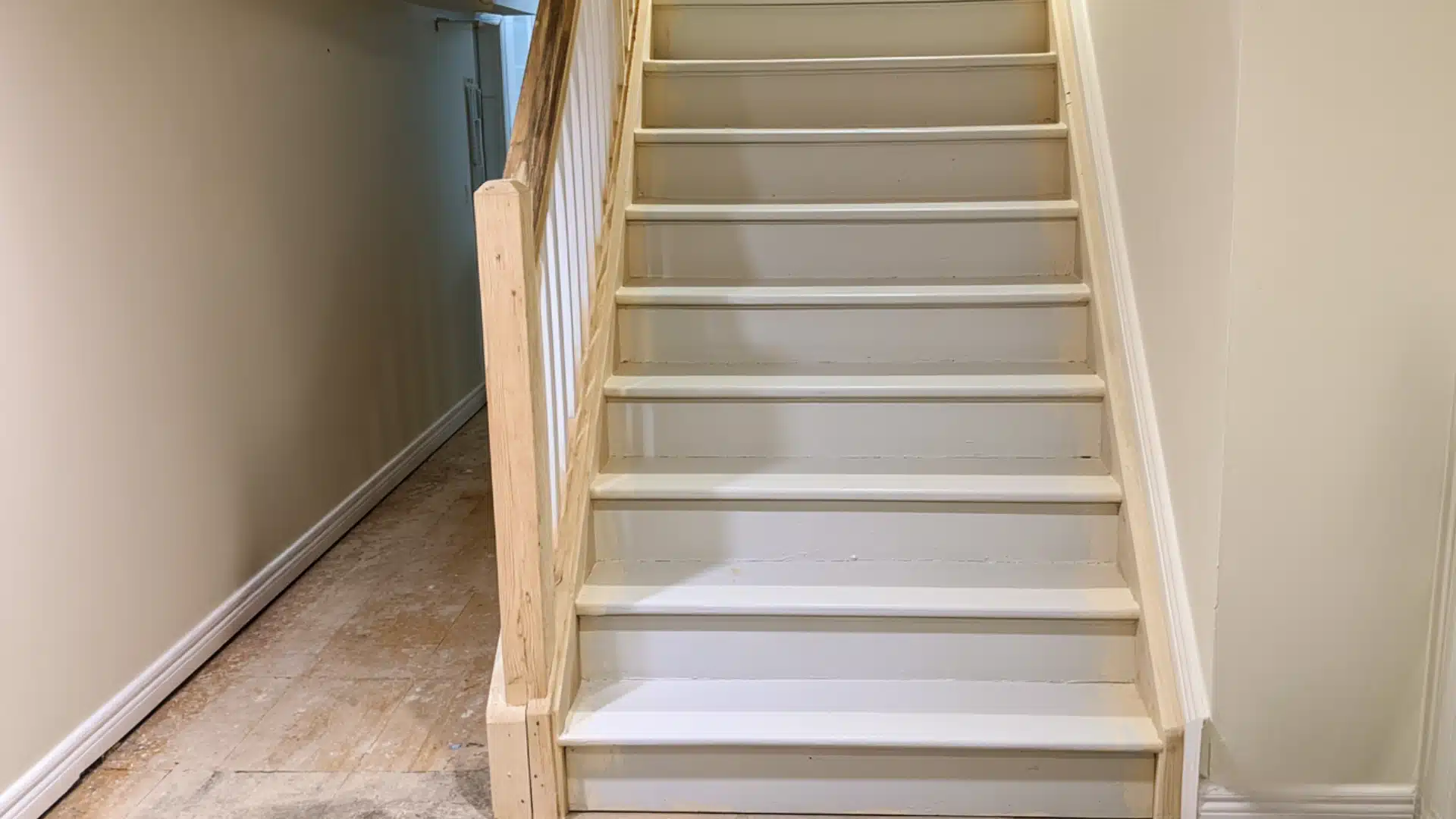 White painted basement staircase with wood handrail leading upstairs beside a narrow unfinished hallway and wood floor