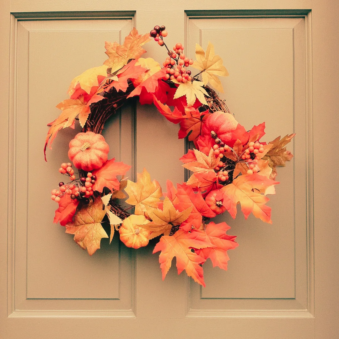 DIY fall wreath with orange maple leaves, mini pumpkins, berries, and grapevine details hanging on a light paneled front door.